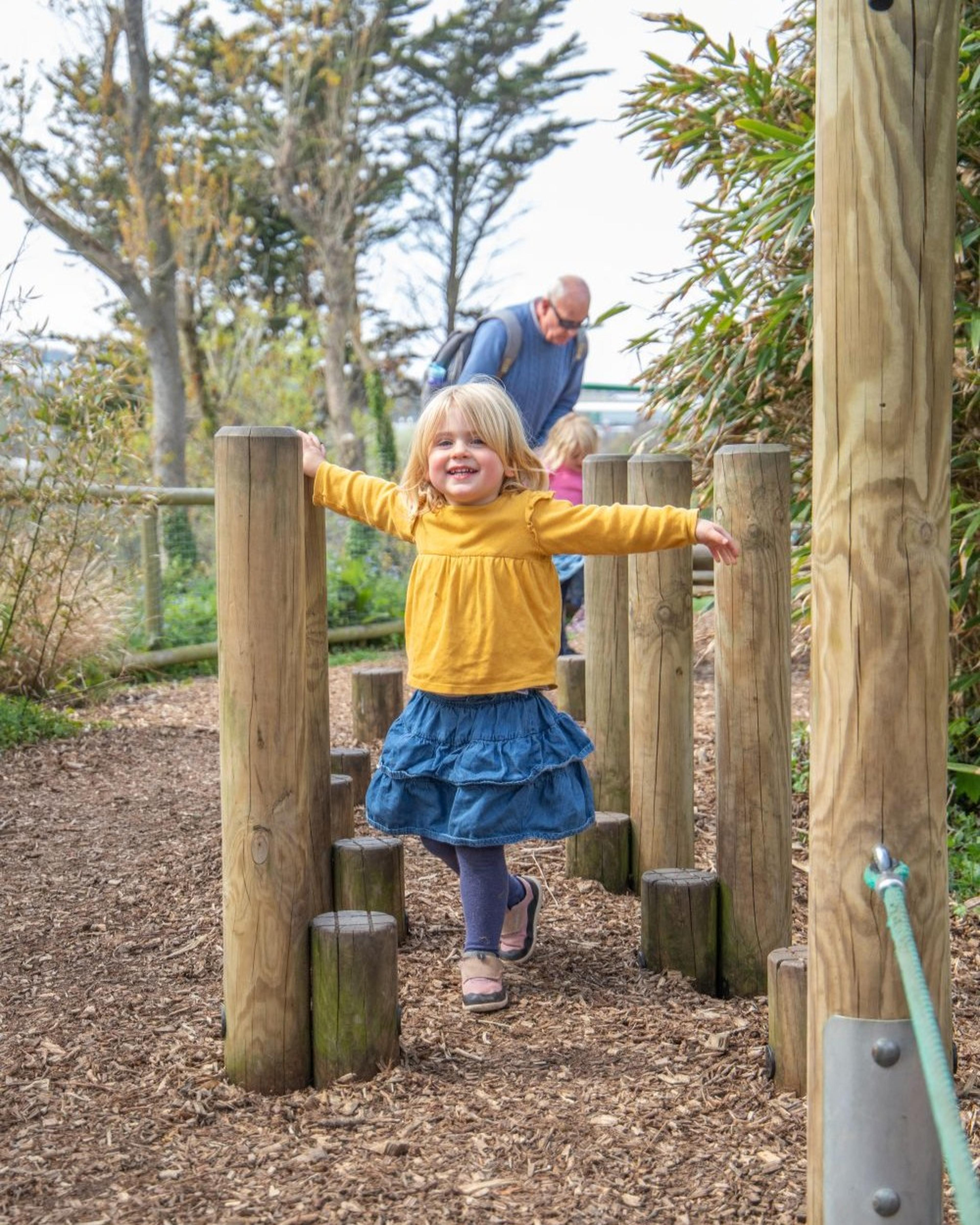 Child in a yellow top and blue skirt balances on wooden posts in a park, arms outstretched, with adults and greenery in the background.