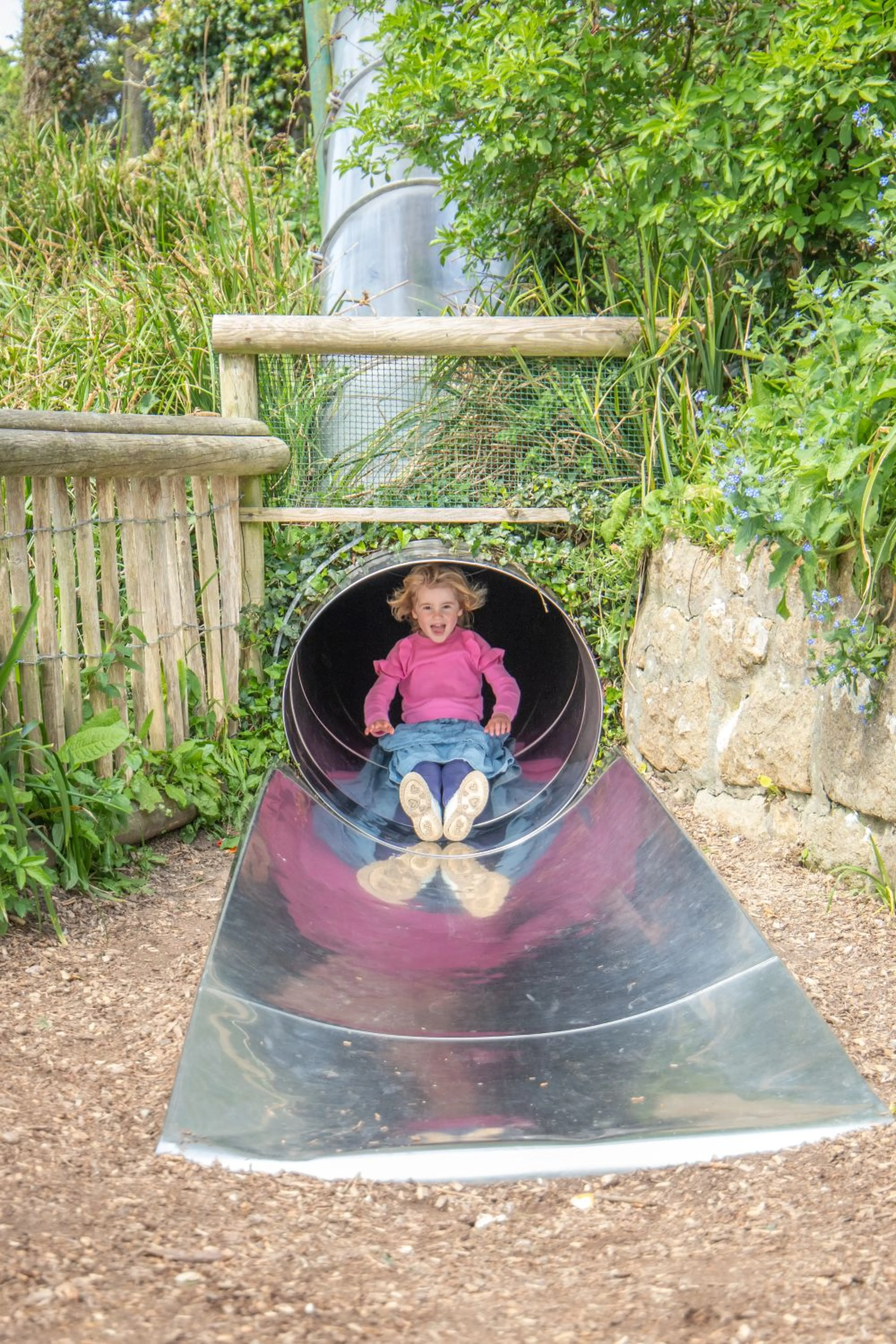Child in pink sweater sliding down a shiny metal tube slide, surrounded by greenery and wooden structures.