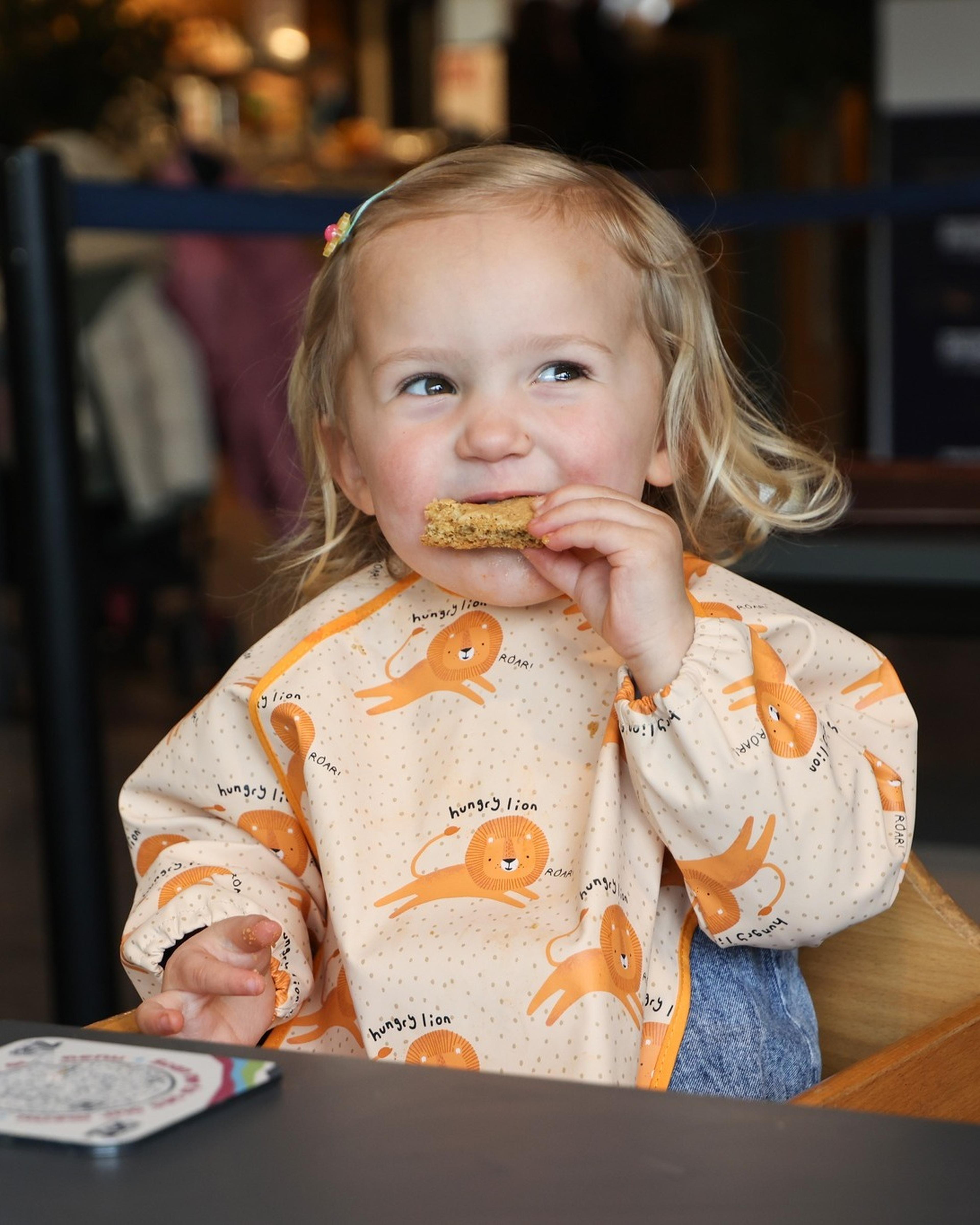 A toddler wearing a bib with playful lion prints sits at a table, smiling and eating a piece of toast.