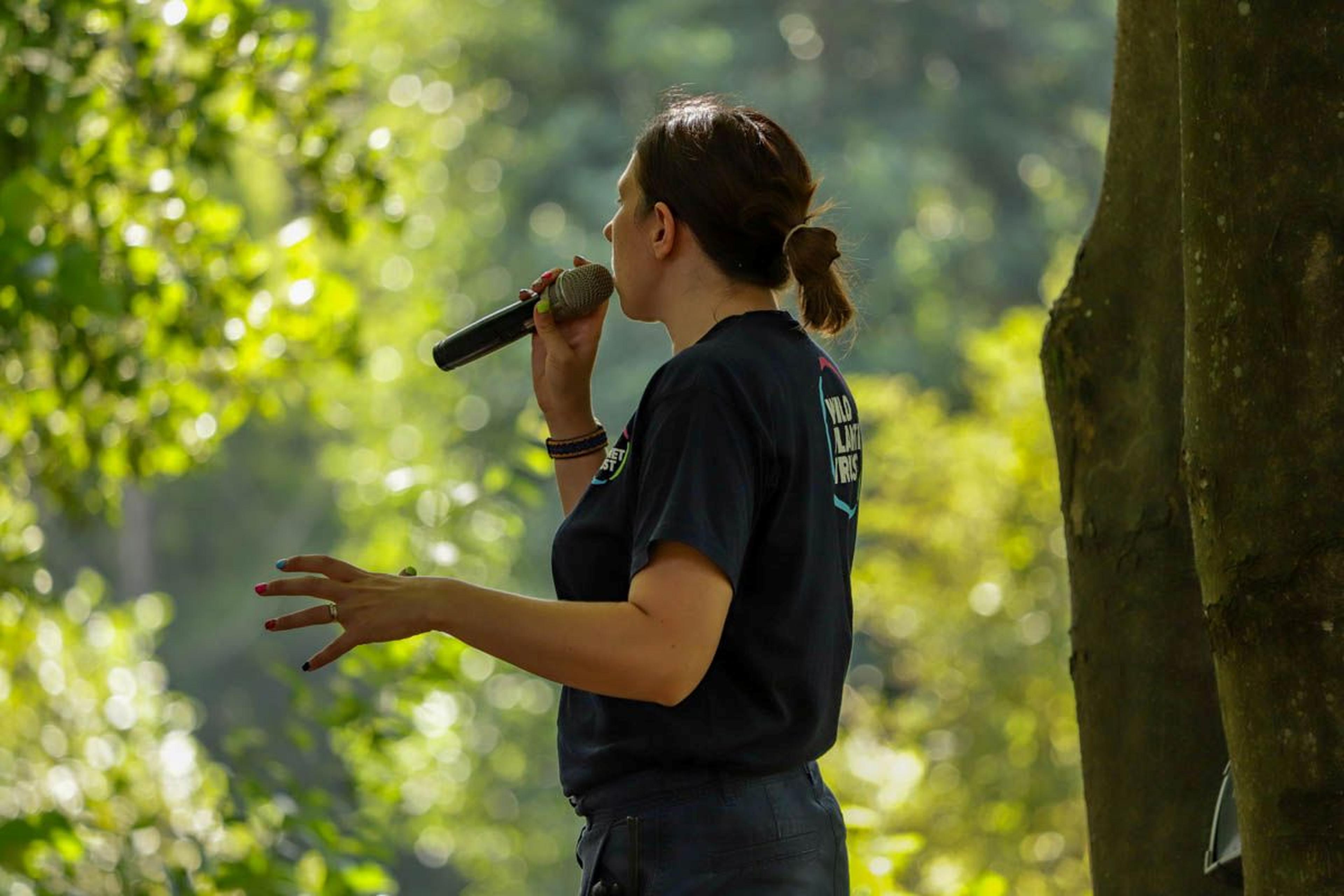 Woman in a black shirt speaking into a microphone outdoors, surrounded by lush green trees.