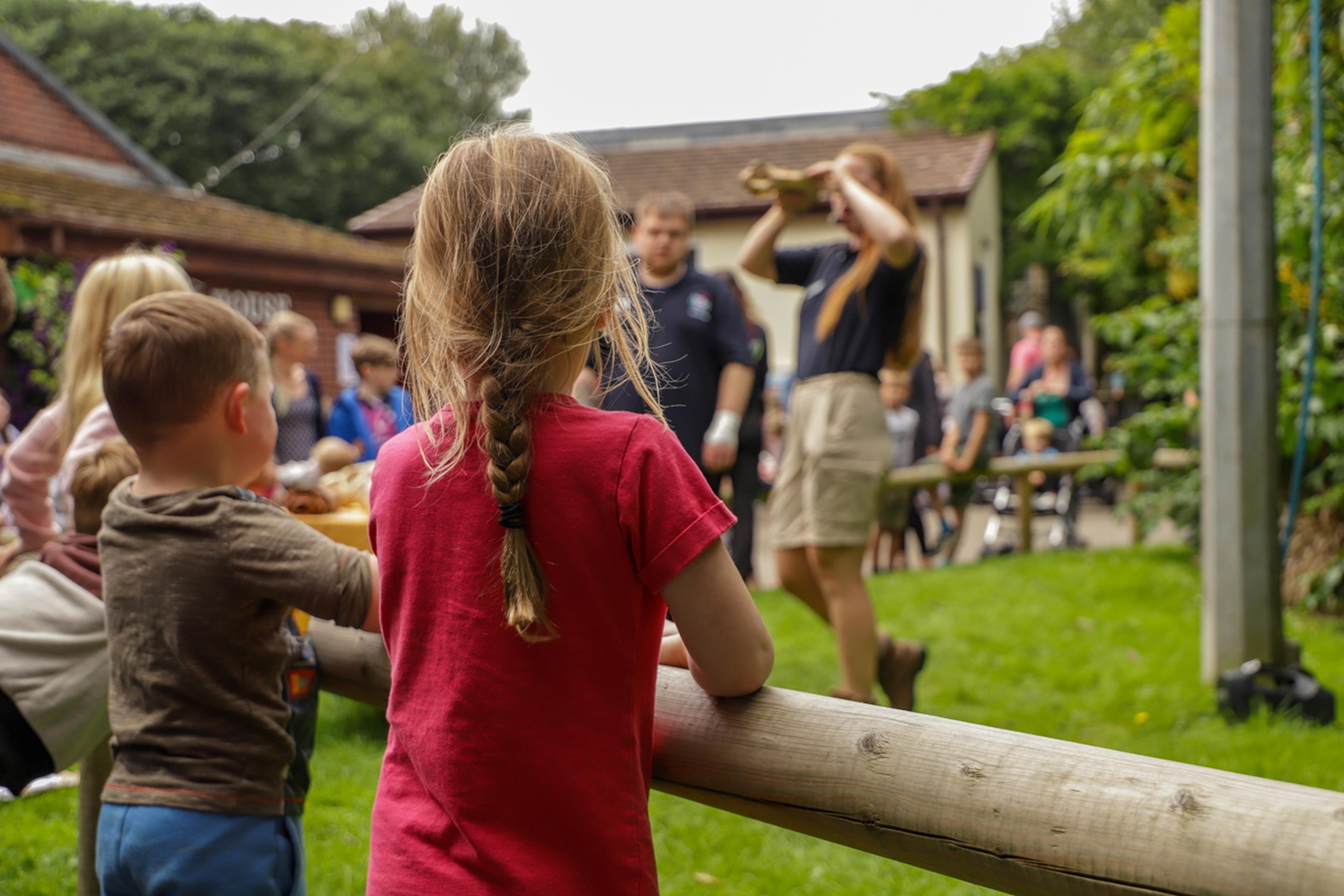 Children watching a zookeeper give a presentation in an outdoor setting, with lush greenery and buildings in the background.