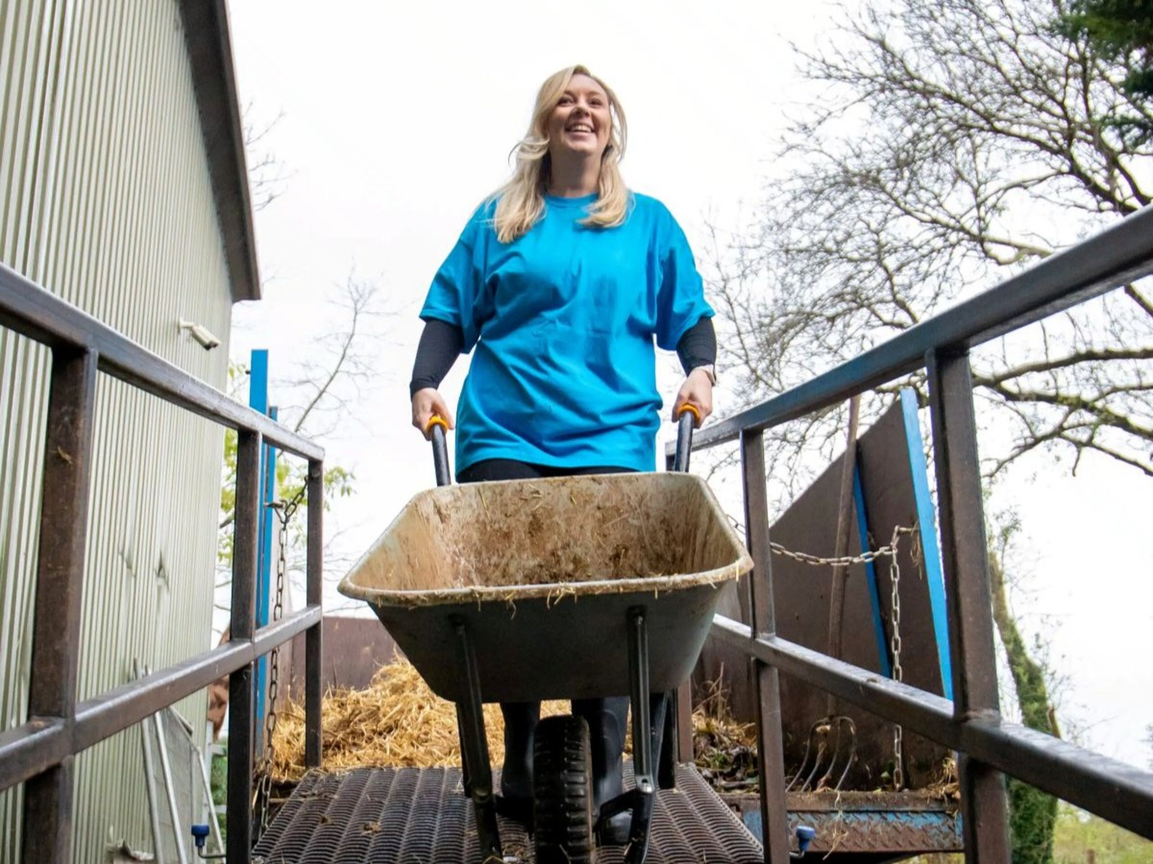 Woman in blue shirt pushing a wheelbarrow filled with hay on a metal ramp, surrounded by trees and a building.