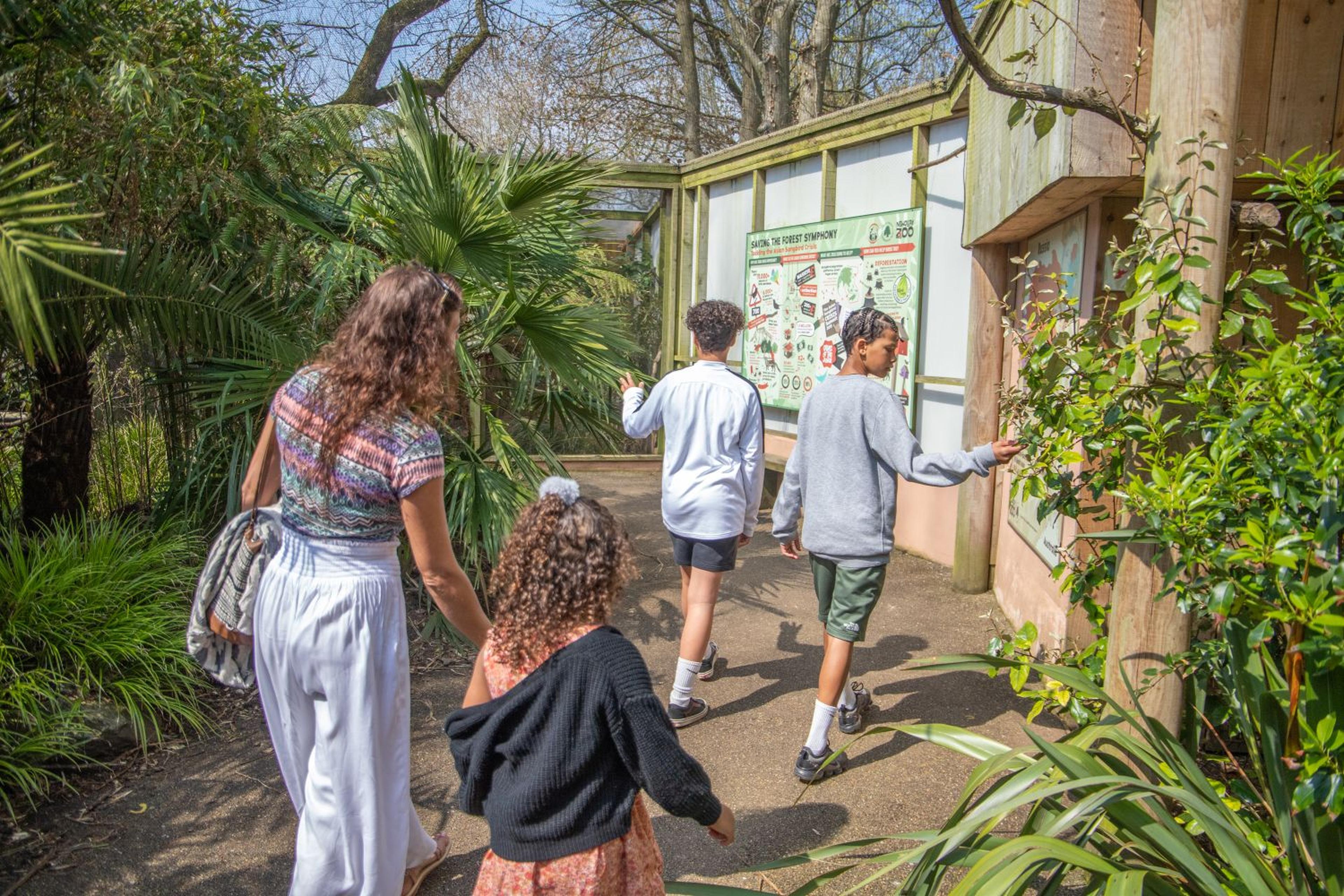 A family walks through a zoo with lush greenery, heading towards an exhibit sign. Bright, sunny day with casual attire.