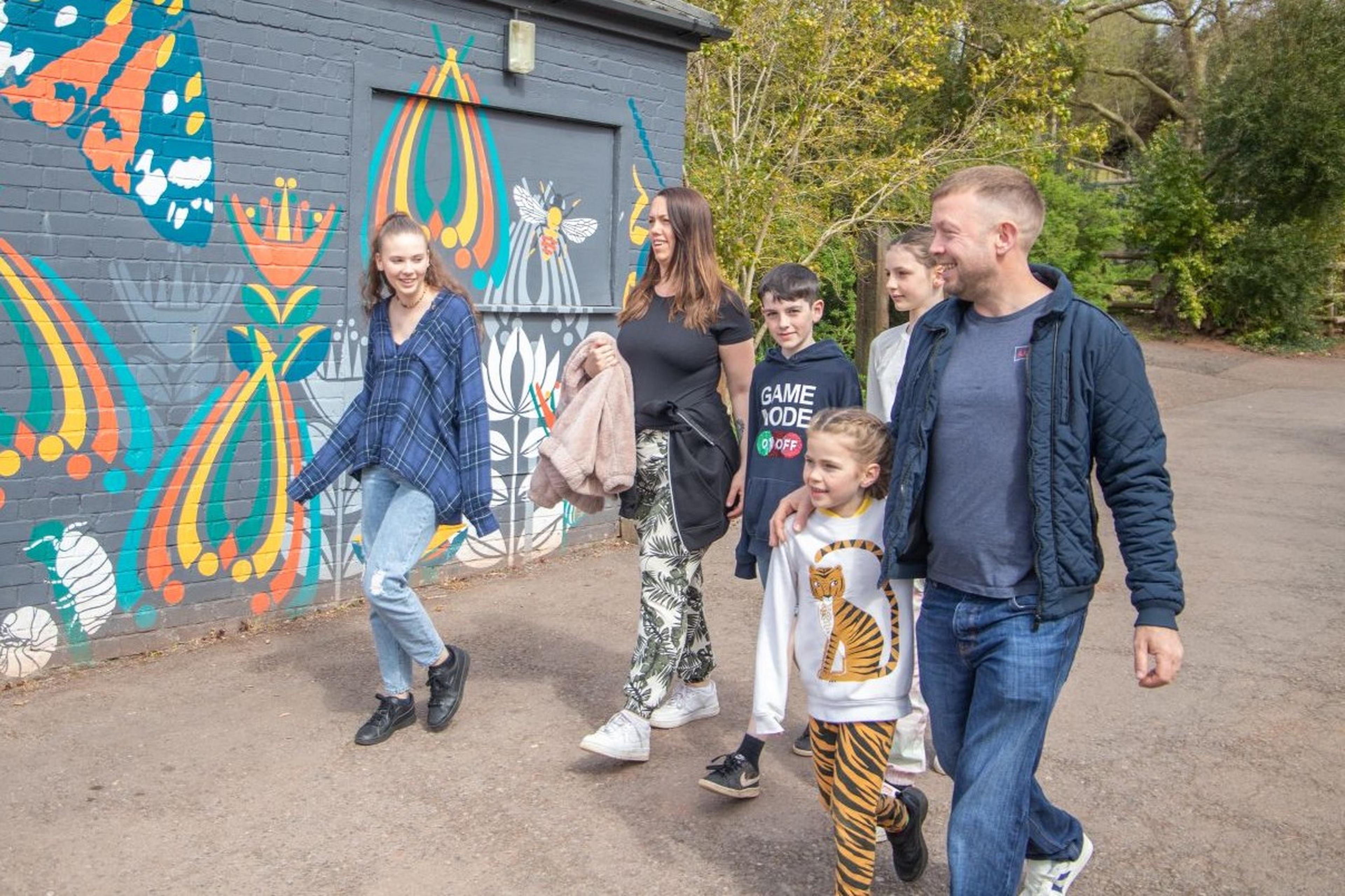 A group of people walking along a path with colorful mural art on a brick wall, featuring butterflies and abstract patterns in the background.