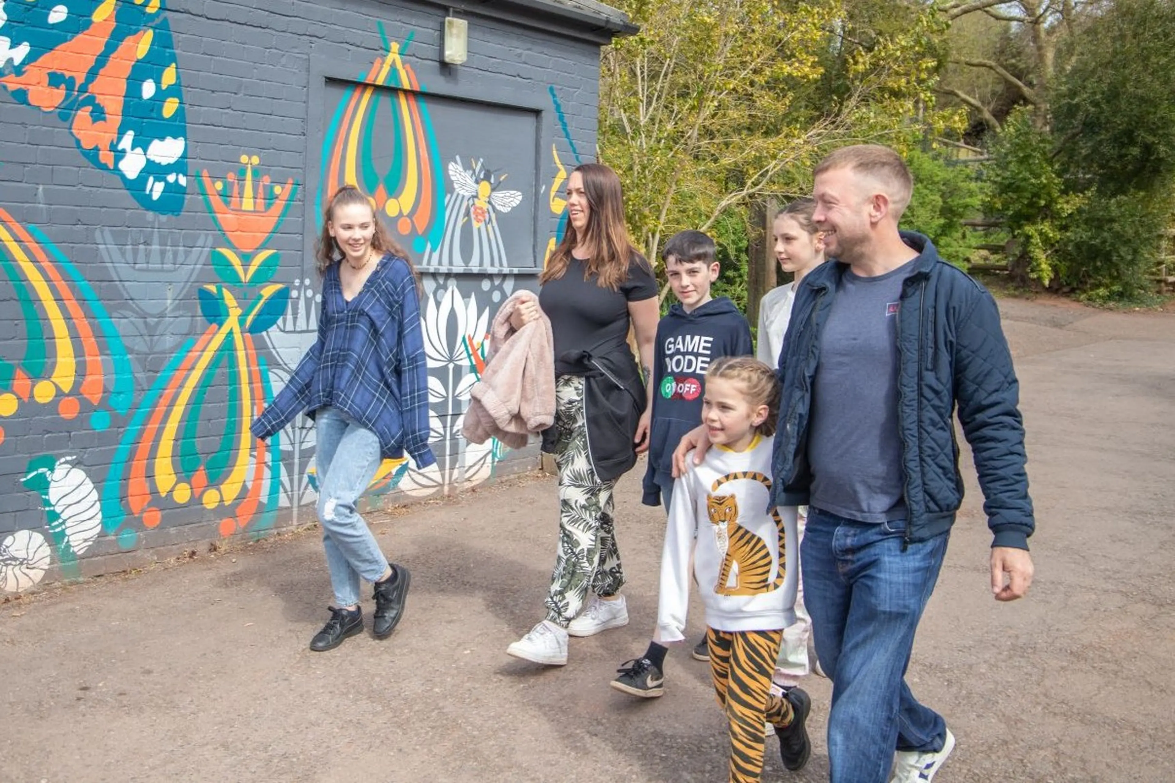 A group of people walking along a path with colorful mural art on a brick wall, featuring butterflies and abstract patterns in the background.