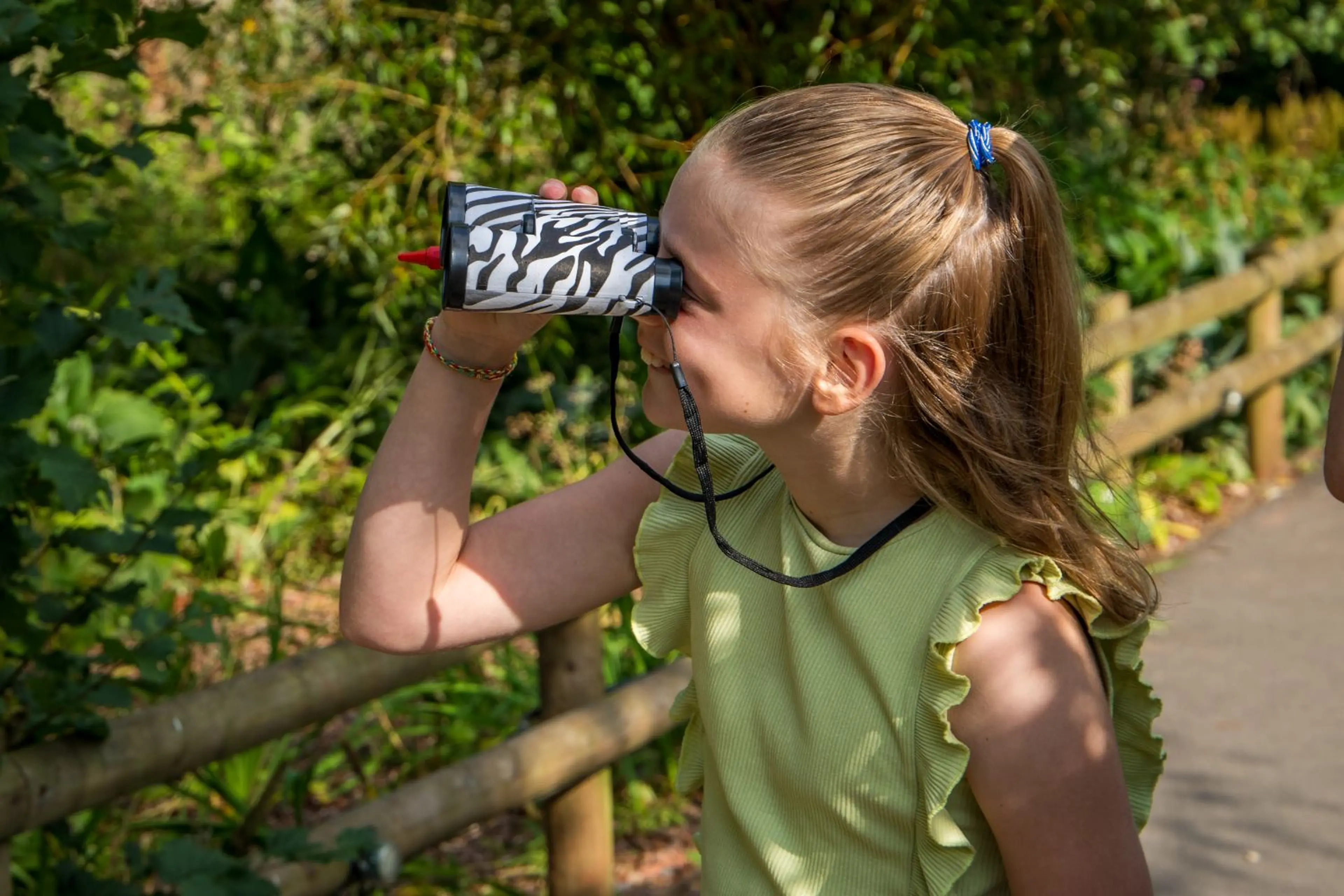 Girl with a ponytail looks through zebra-print binoculars in a garden, wearing a sleeveless green top. Sunlight filters through surrounding trees.