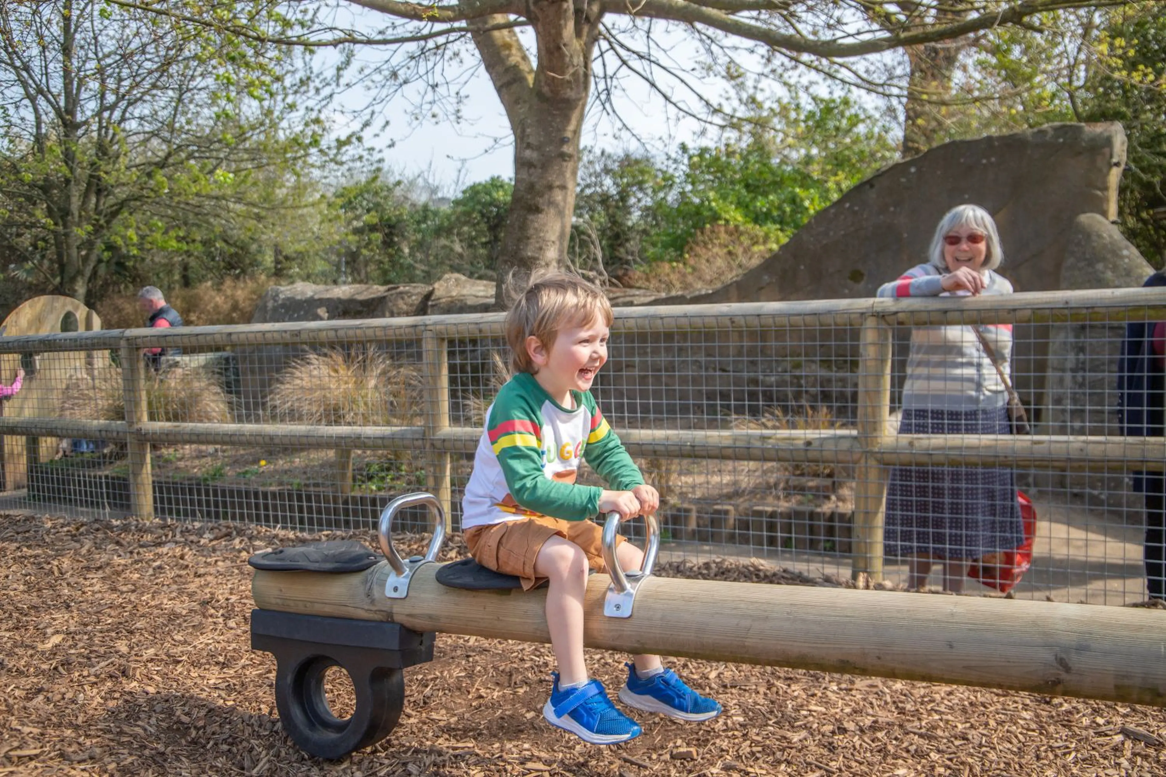 A young boy is joyfully riding a seesaw outdoors, under the watchful eye of a smiling woman standing nearby, with trees in the background.