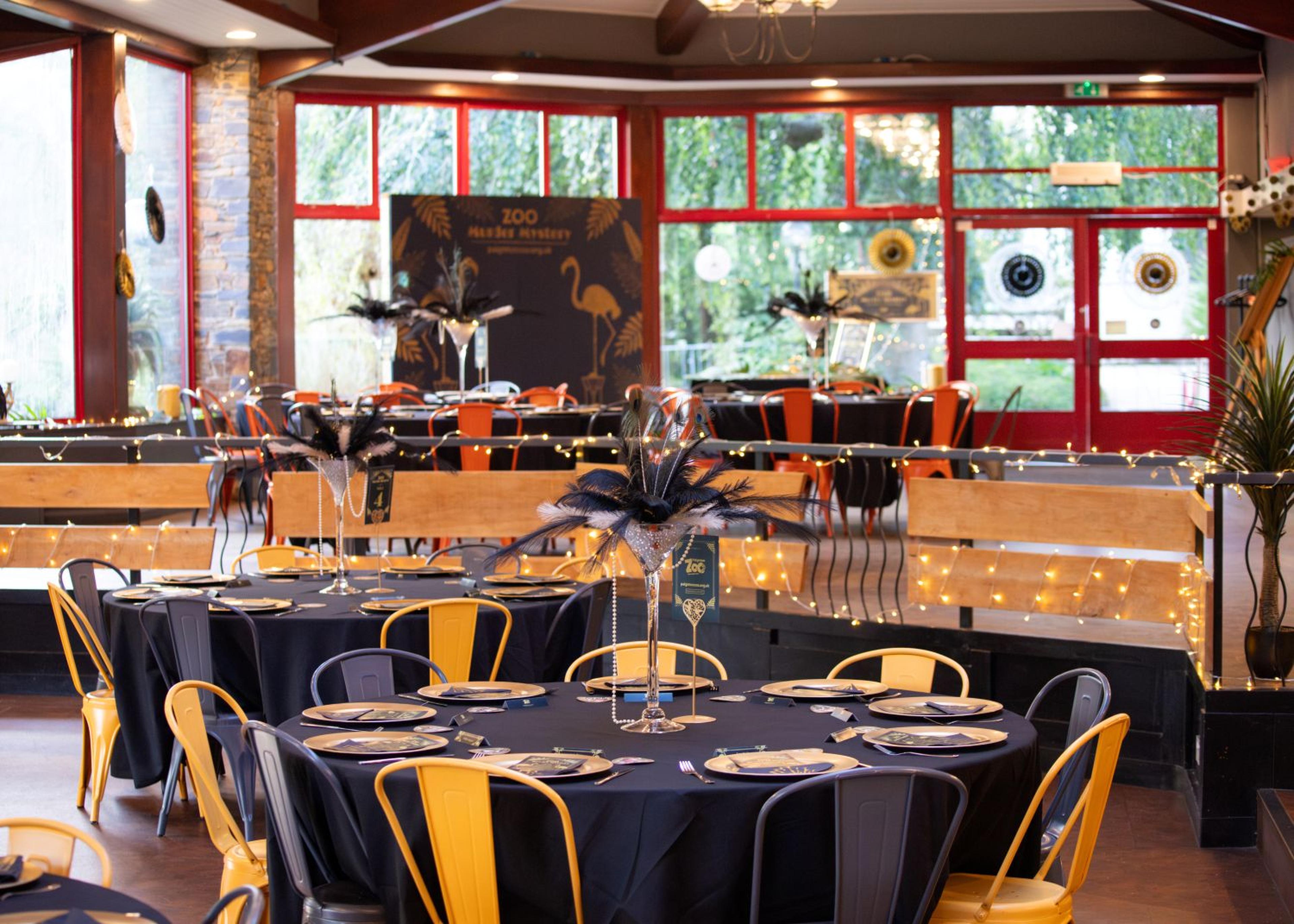 Elegant event venue with round tables, black tablecloths, and feather centerpieces. Warm lighting and large windows create an inviting atmosphere.
