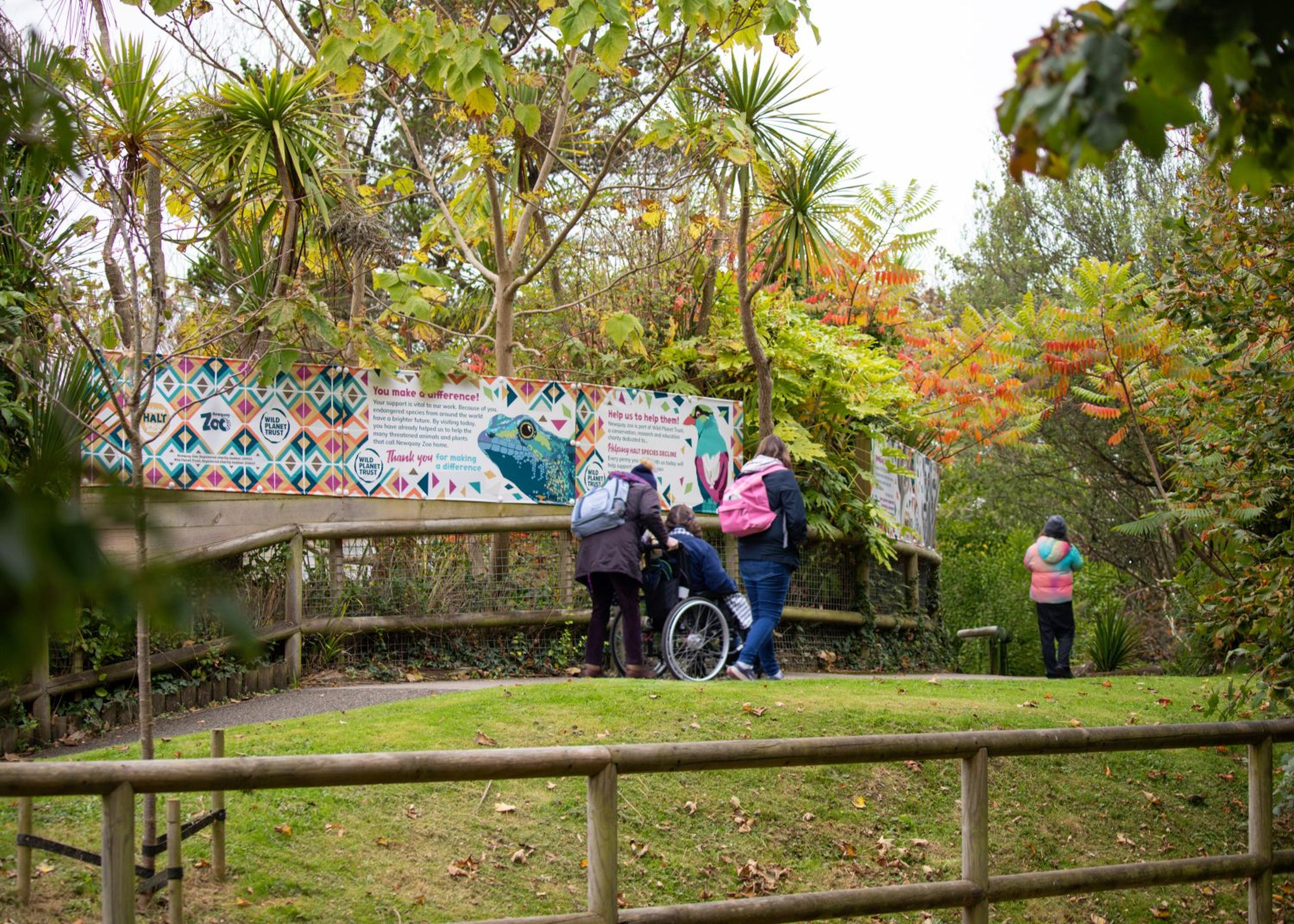 People stand near a colorful mural on an outdoor path, surrounded by lush greenery and trees, including palm and autumn-colored leaves.