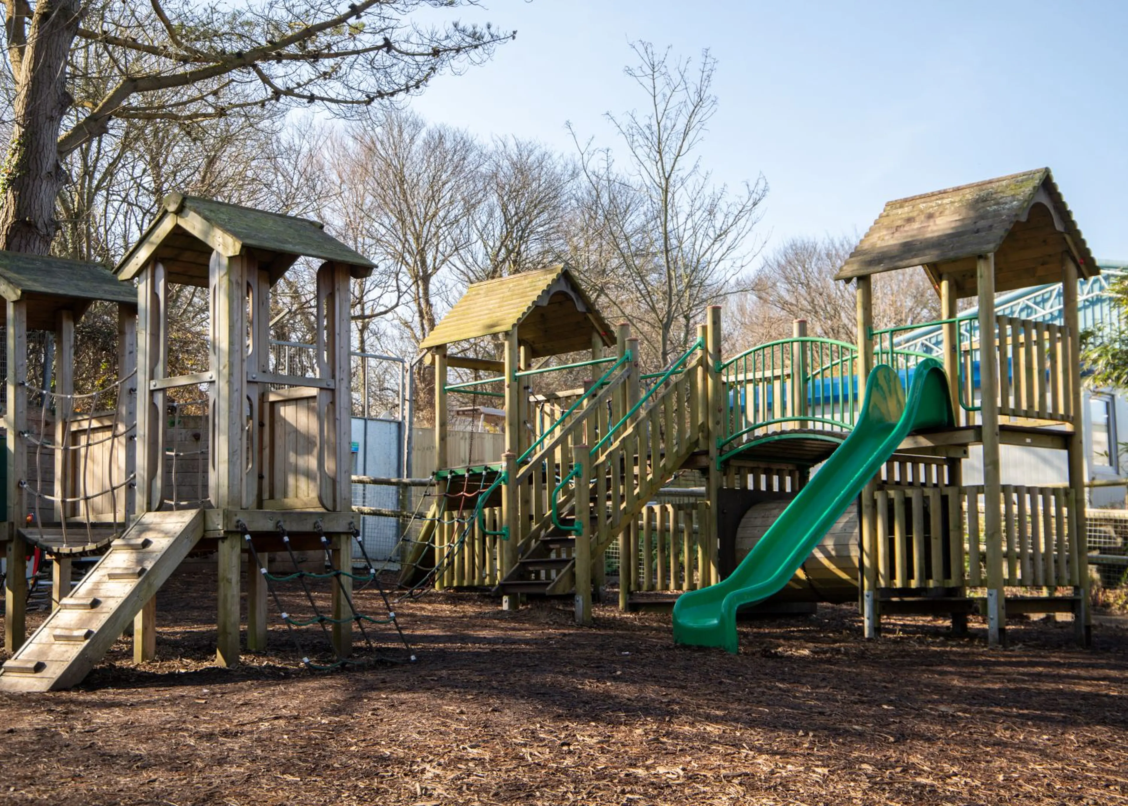 Wooden playground with slide, rope ladder, and bridges under clear skies, surrounded by trees with bare branches.
