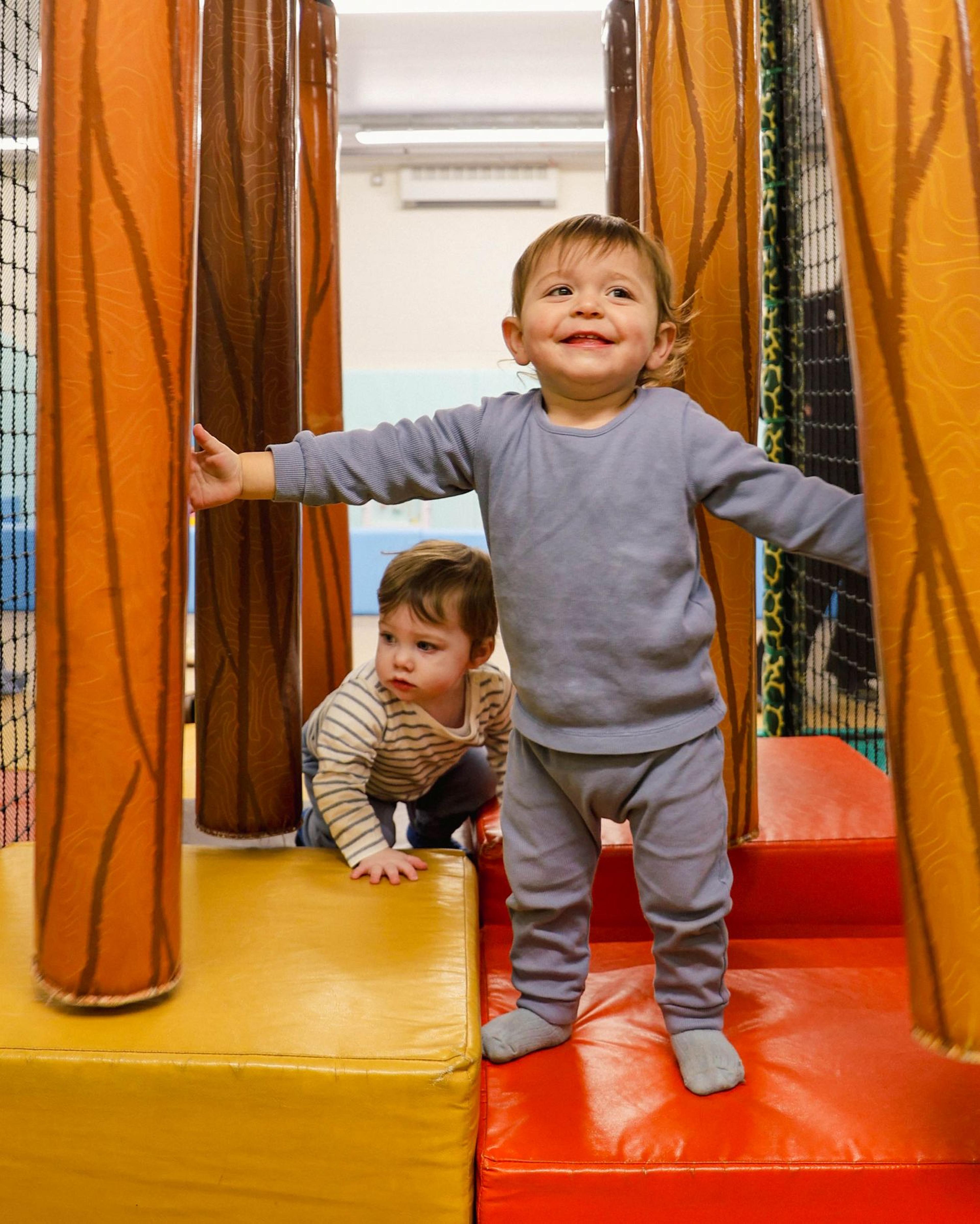 Two toddlers play on a padded indoor playground. One stands and smiles, while the other crouches on the floor, surrounded by tree-like structures.