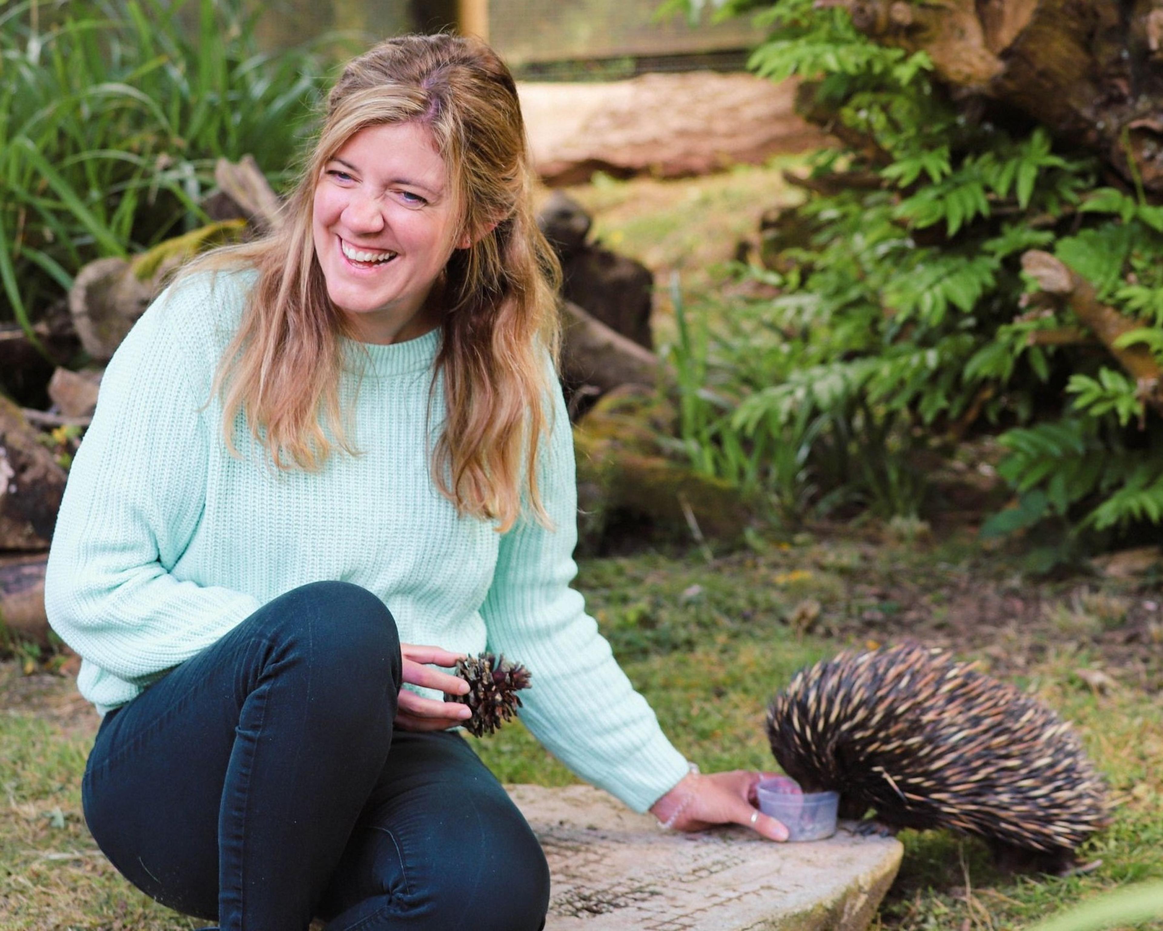 Smiling woman in light sweater sits outdoors on a log, holding a pinecone, with an echidna nearby on grass surrounded by greenery.