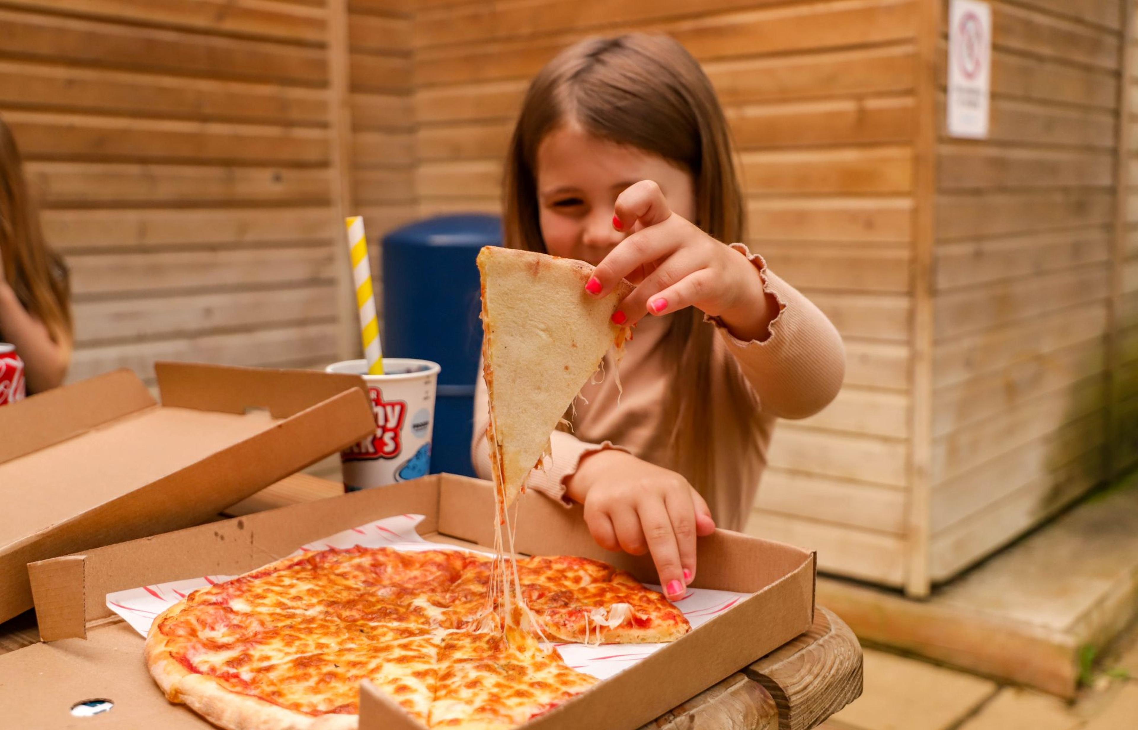A young girl pulls a slice of cheese pizza from a box on a wooden table outside. A drink with a striped straw is beside her.