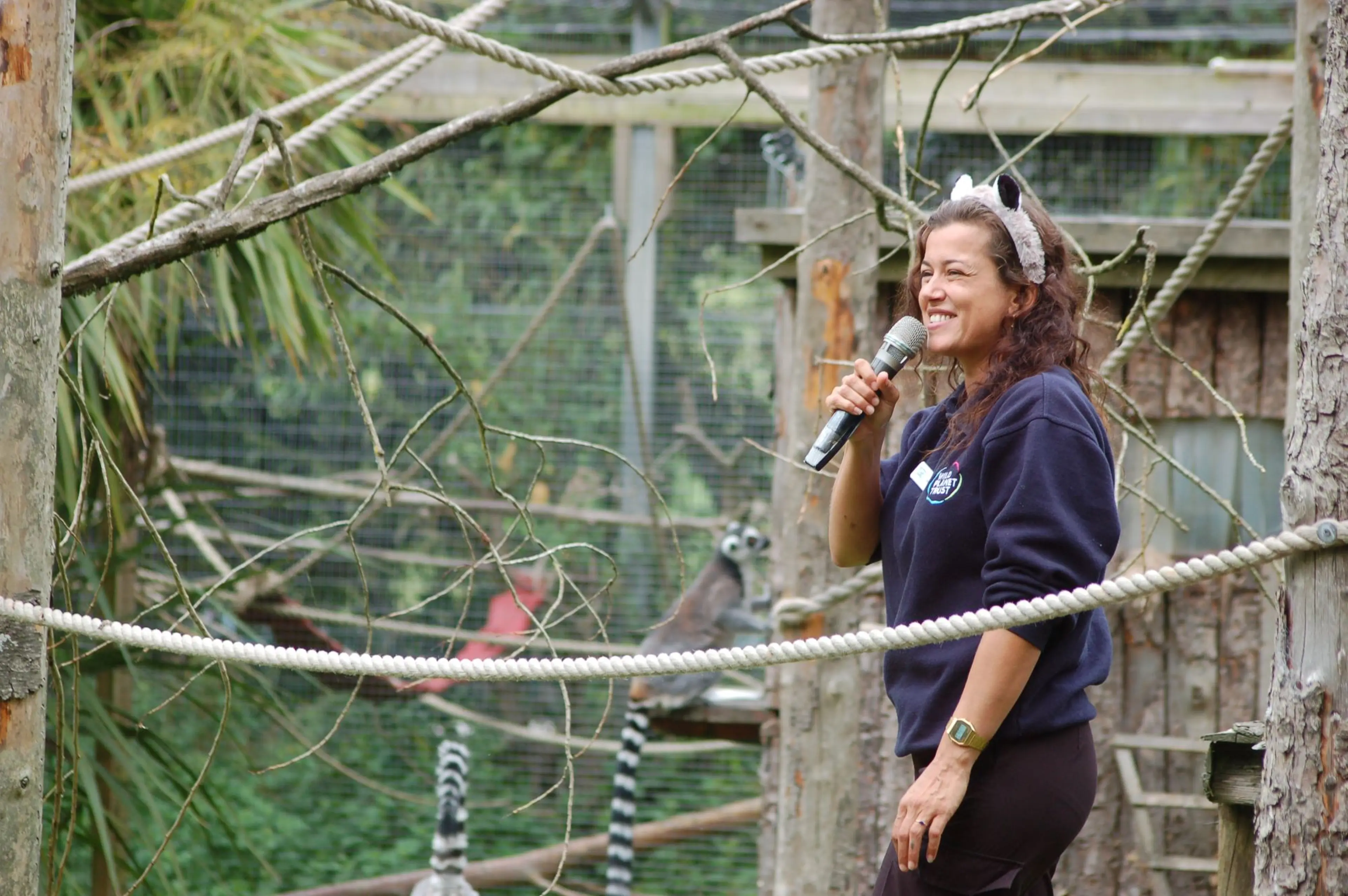 Woman with animal ears headband speaking into a microphone in a zoo enclosure, with lemurs in the background on ropes.