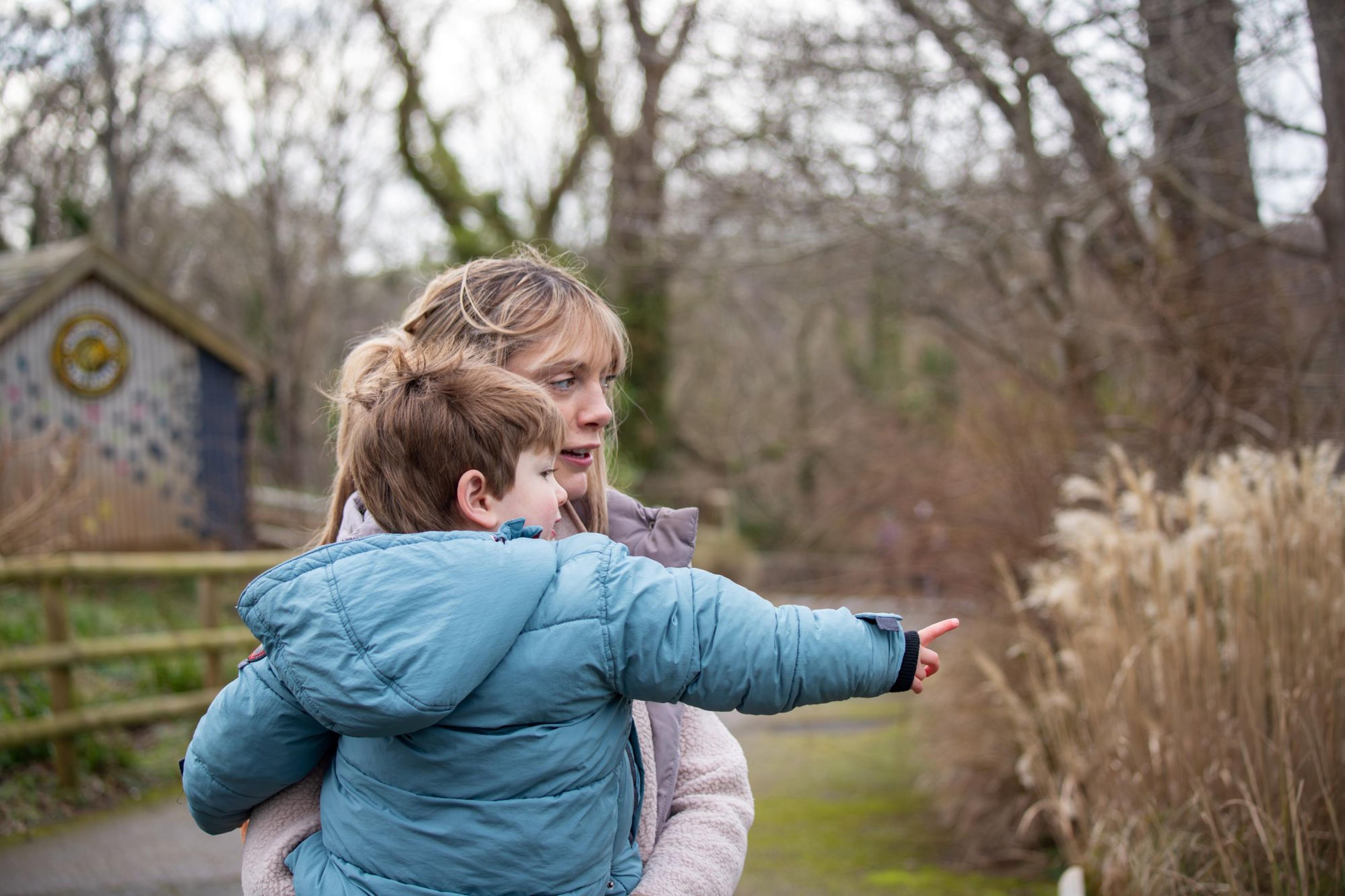 Woman holding a child pointing towards bare trees and dry grasses on a cloudy day in a park.