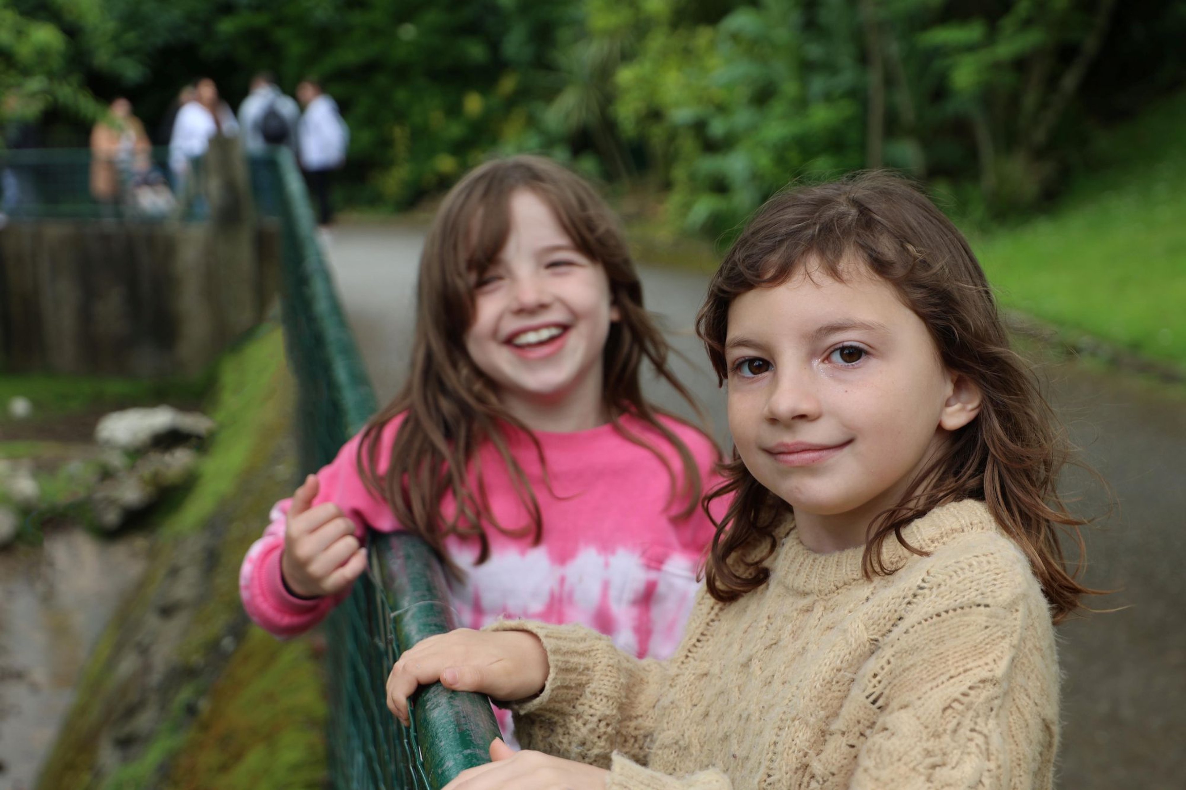 Two young girls stand by a railing in a park. One wears a pink tie-dye shirt, the other a knitted beige sweater. Both are smiling, with greenery around.
