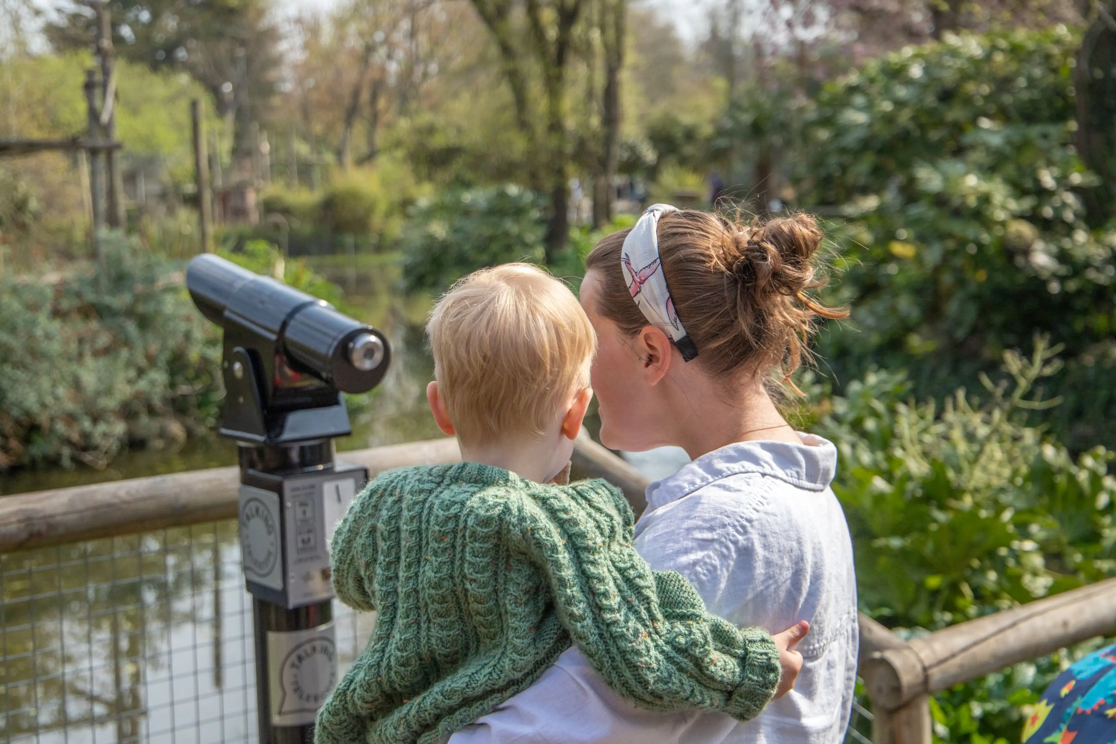 Woman holding a child gazing into a coin-operated viewer at a zoo or park, surrounded by lush greenery on a sunny day.