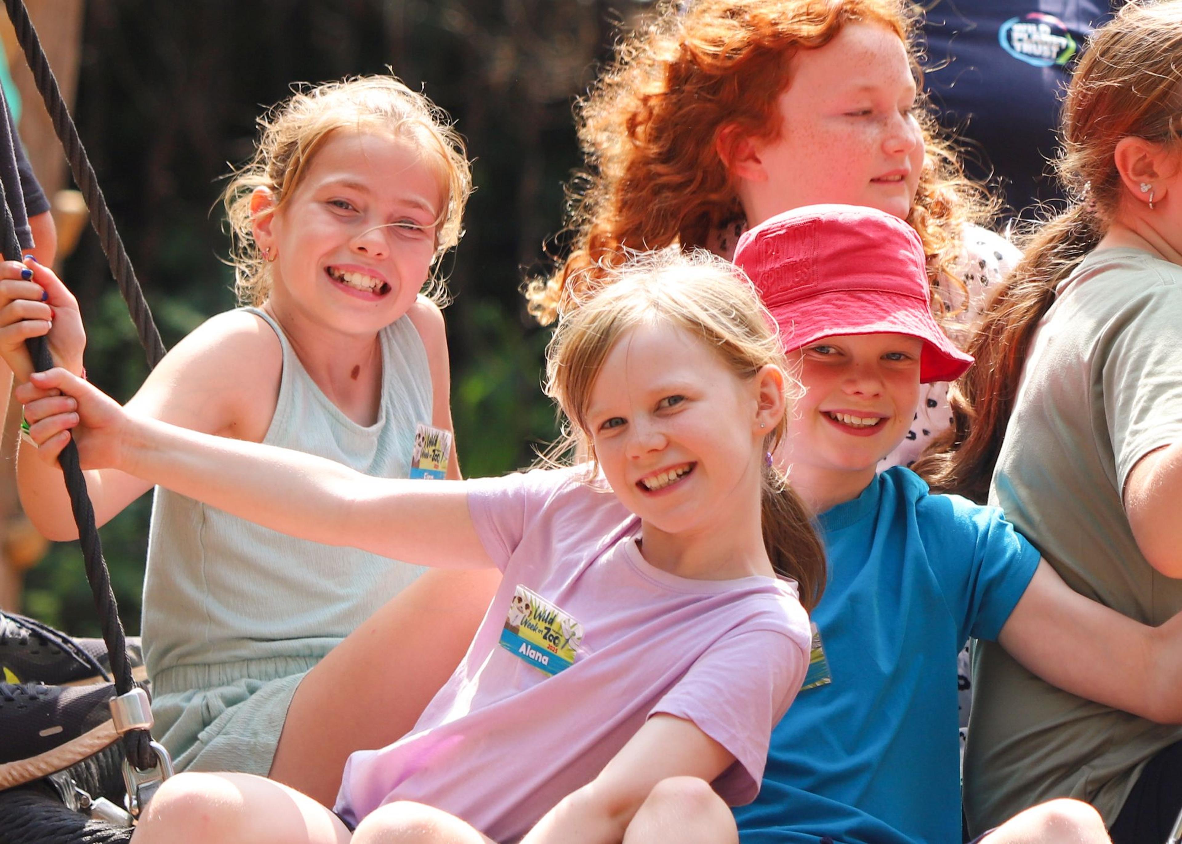 Four smiling children riding a swing outdoors, enjoying a sunny day. One wears a red hat; others have casual summer attire.