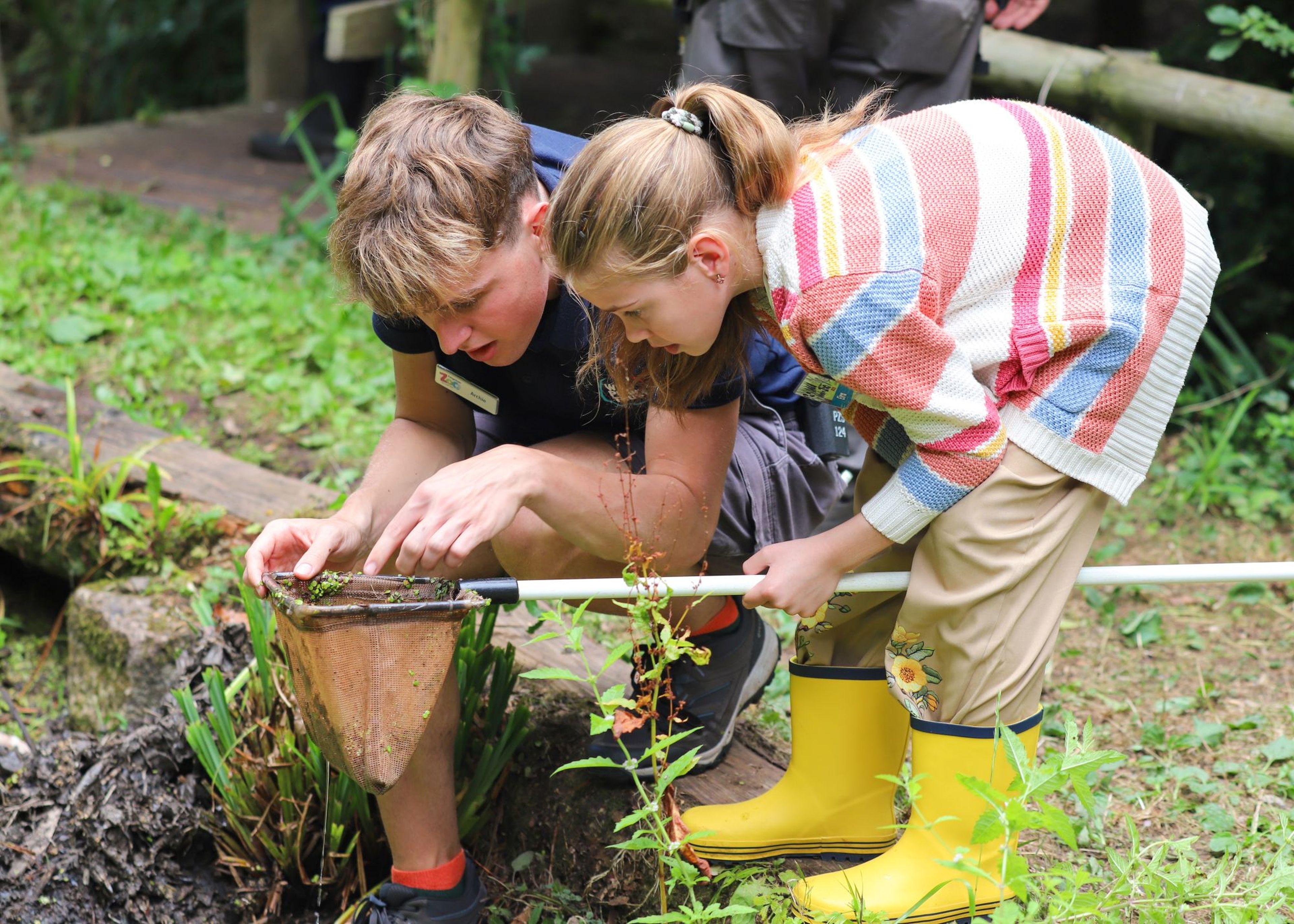 Two children examine a net beside a pond in a garden, both focused closely on the contents. The girl wears yellow boots and a colorful sweater.