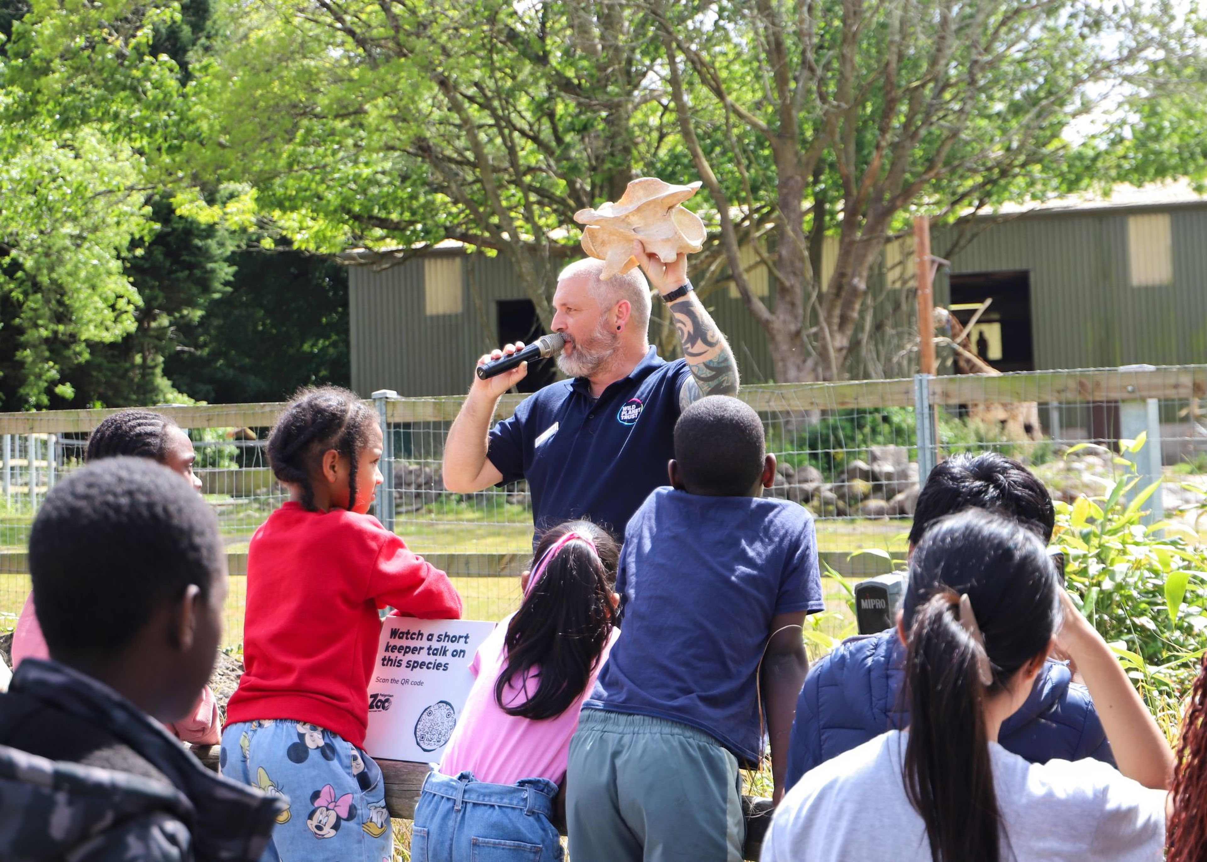 A man holding a large shell speaks into a microphone to a group of children outdoors, near a fence and trees.
