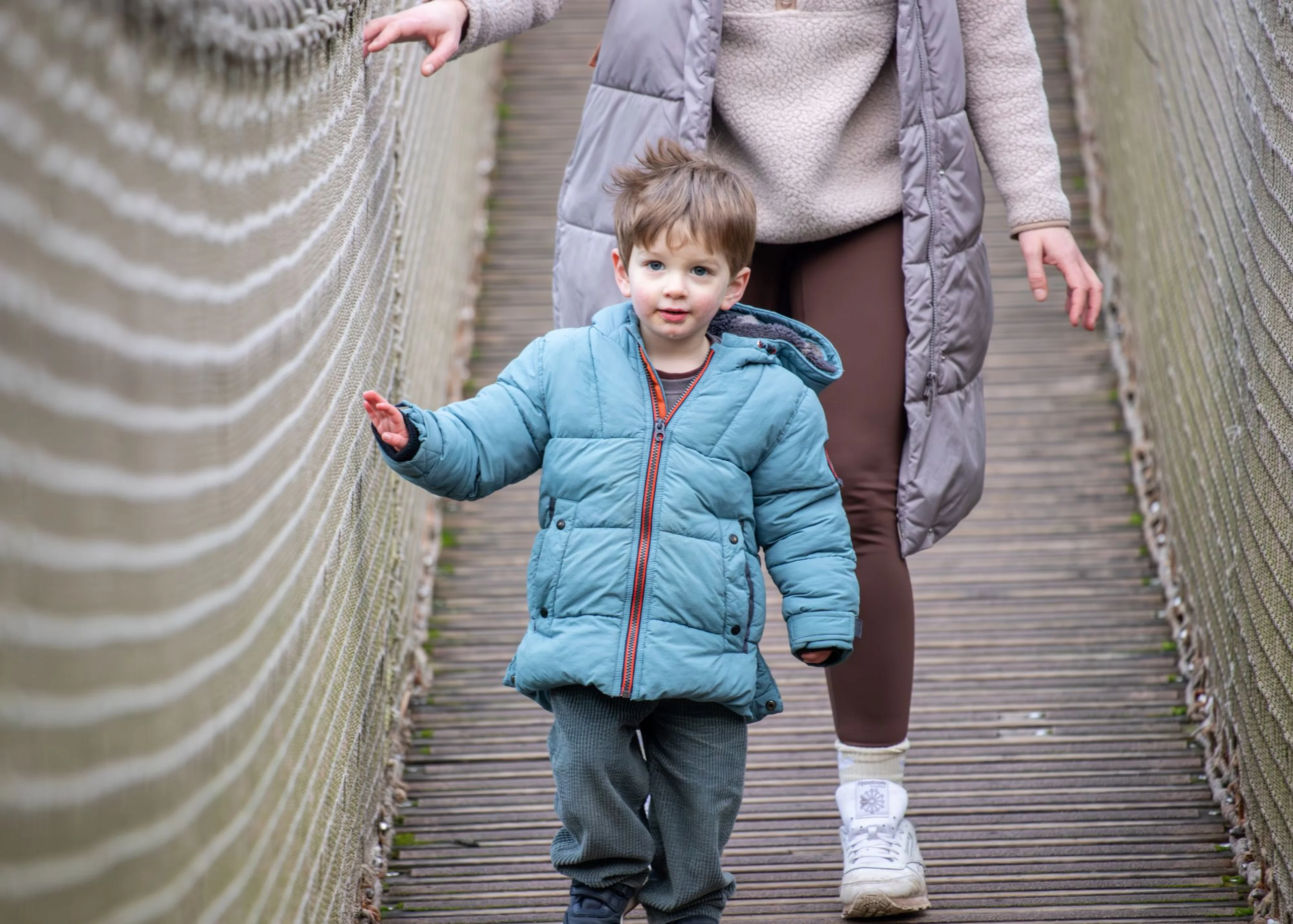 Child in a blue jacket walking on a wooden bridge, followed by an adult in a gray coat and brown pants.