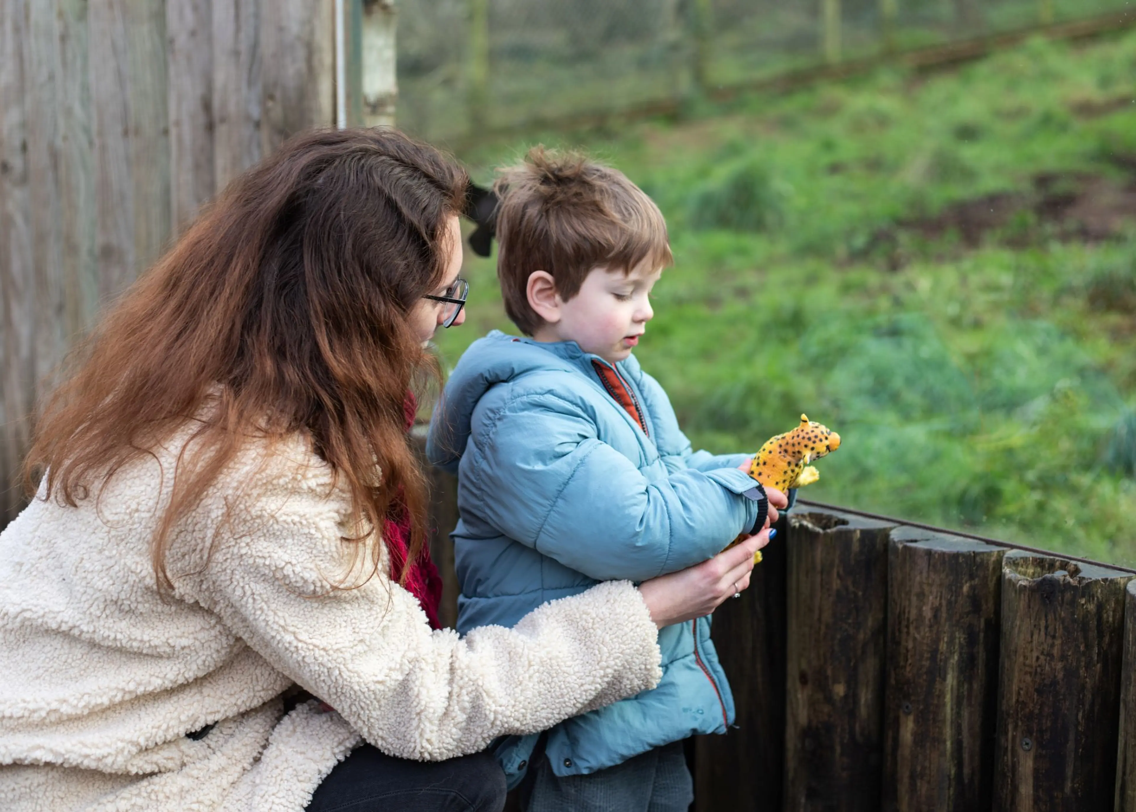Woman and young boy in warm coats observe animals over a wooden fence. The boy holds a small plush giraffe. Green grass in the background.
