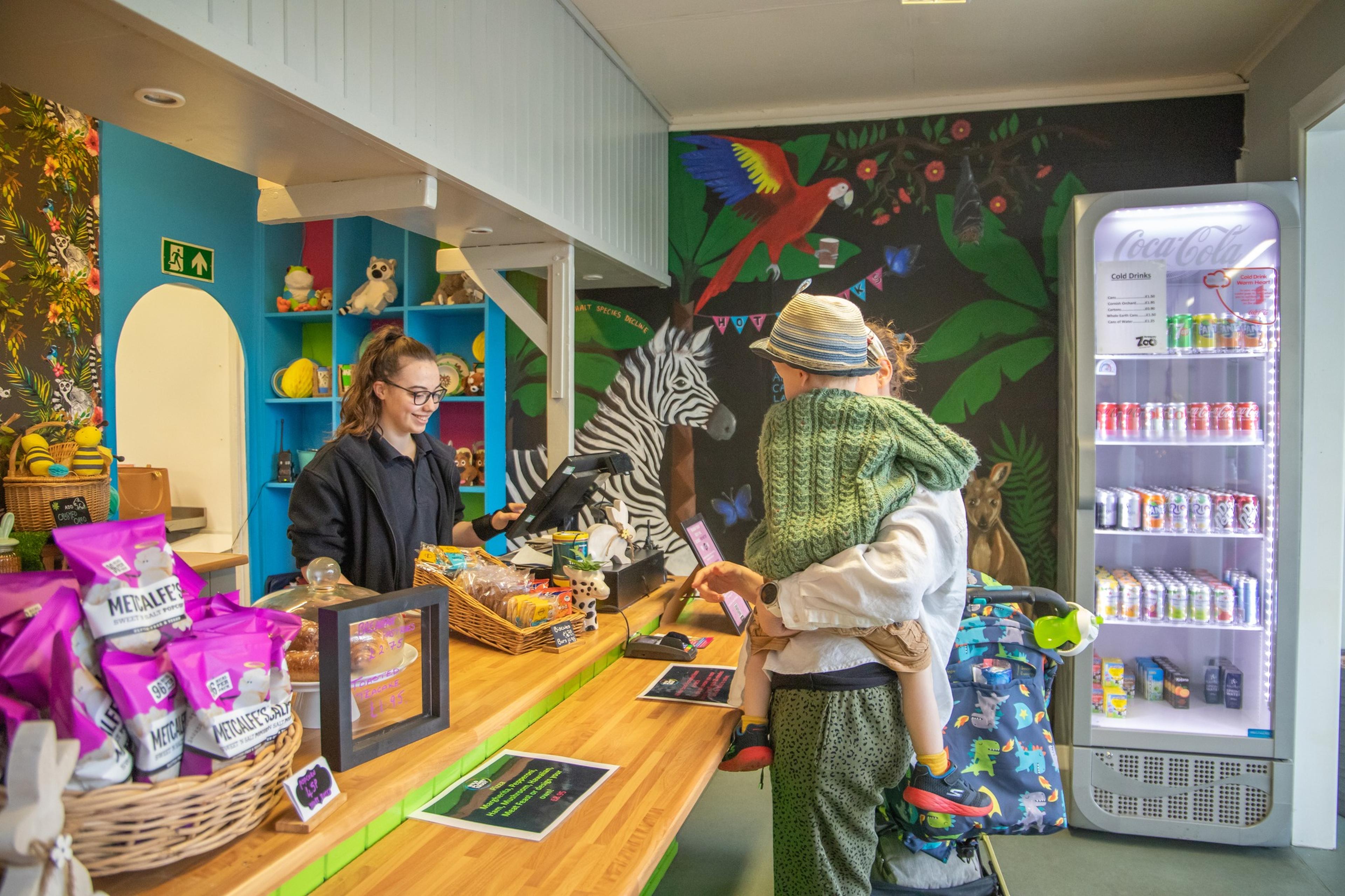 Woman with child at colorful store counter with snacks and drinks, vibrant animal mural, and beverage cooler in the background.