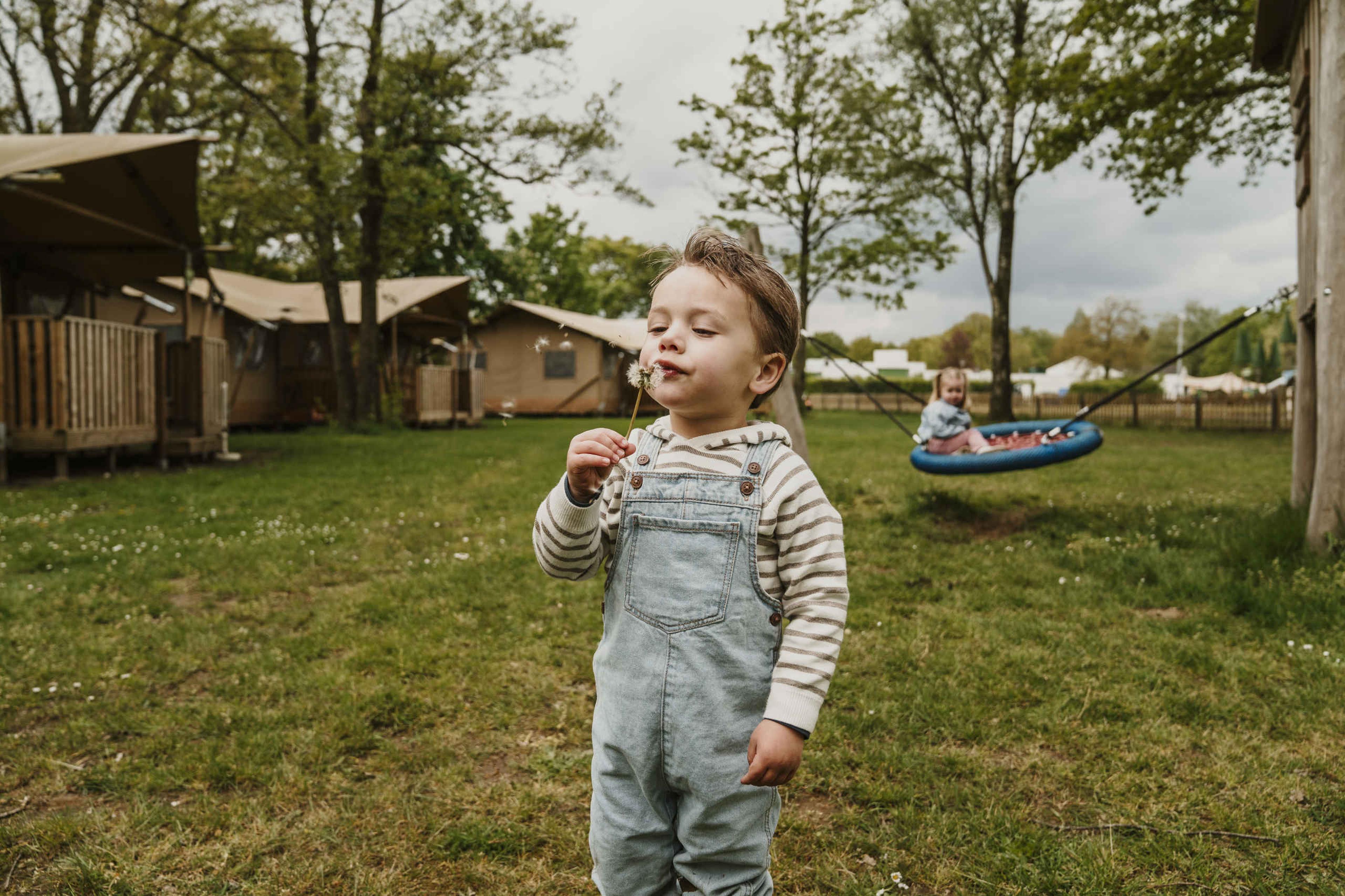 Een jongen blaast een paardenbloem weg en een meisje zit op de schommel bij Vakantiepark Dierenbos