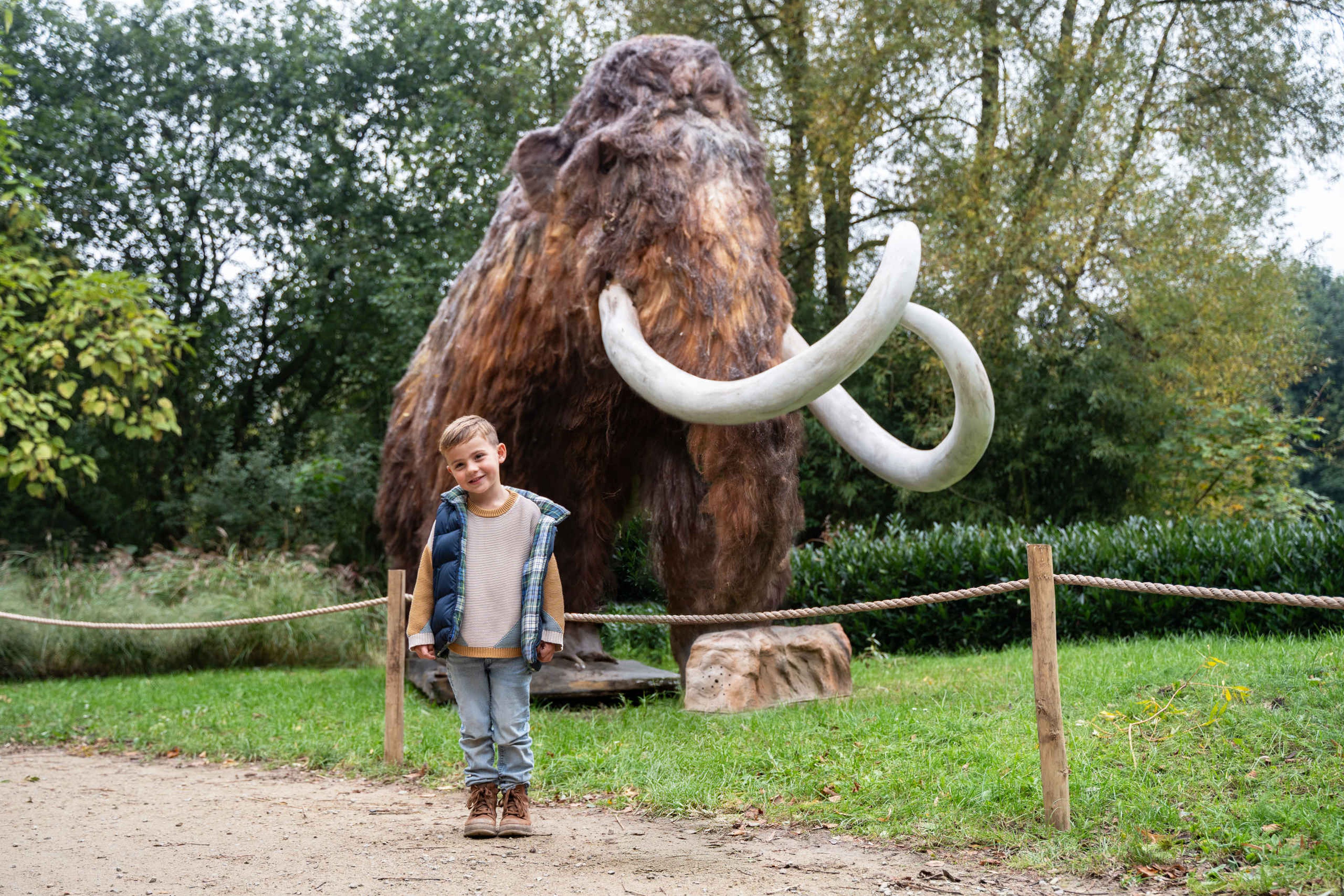 Een jongetje posseert voor de foto met een wolharige mammoet op de achtergrond tijdens IJstijd in ZooParc Overloon