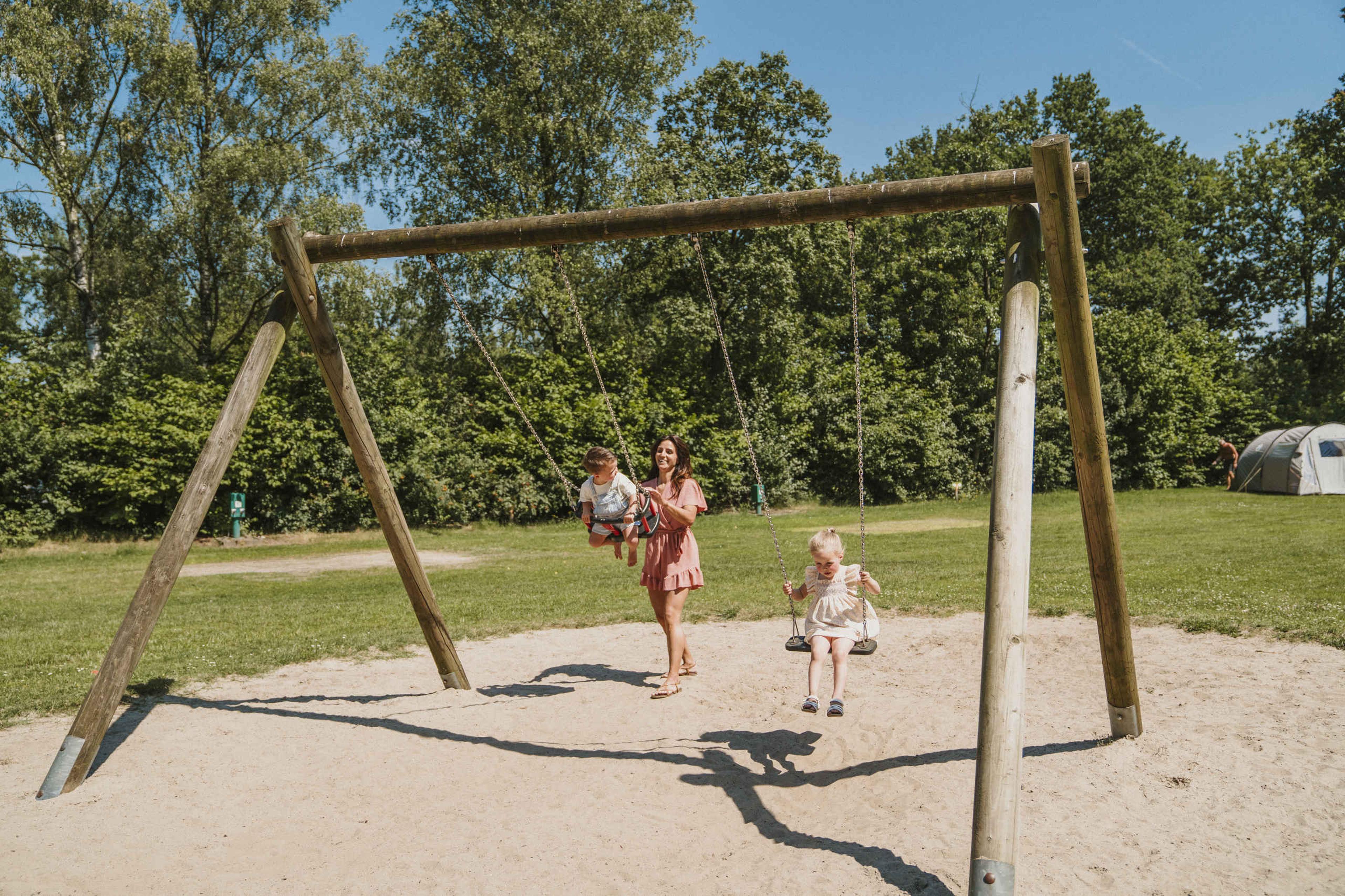 Een moeder schommelt met haar kinderen in de speeltuin op de kampeerplaats bij Vakantiepark Dierenbos