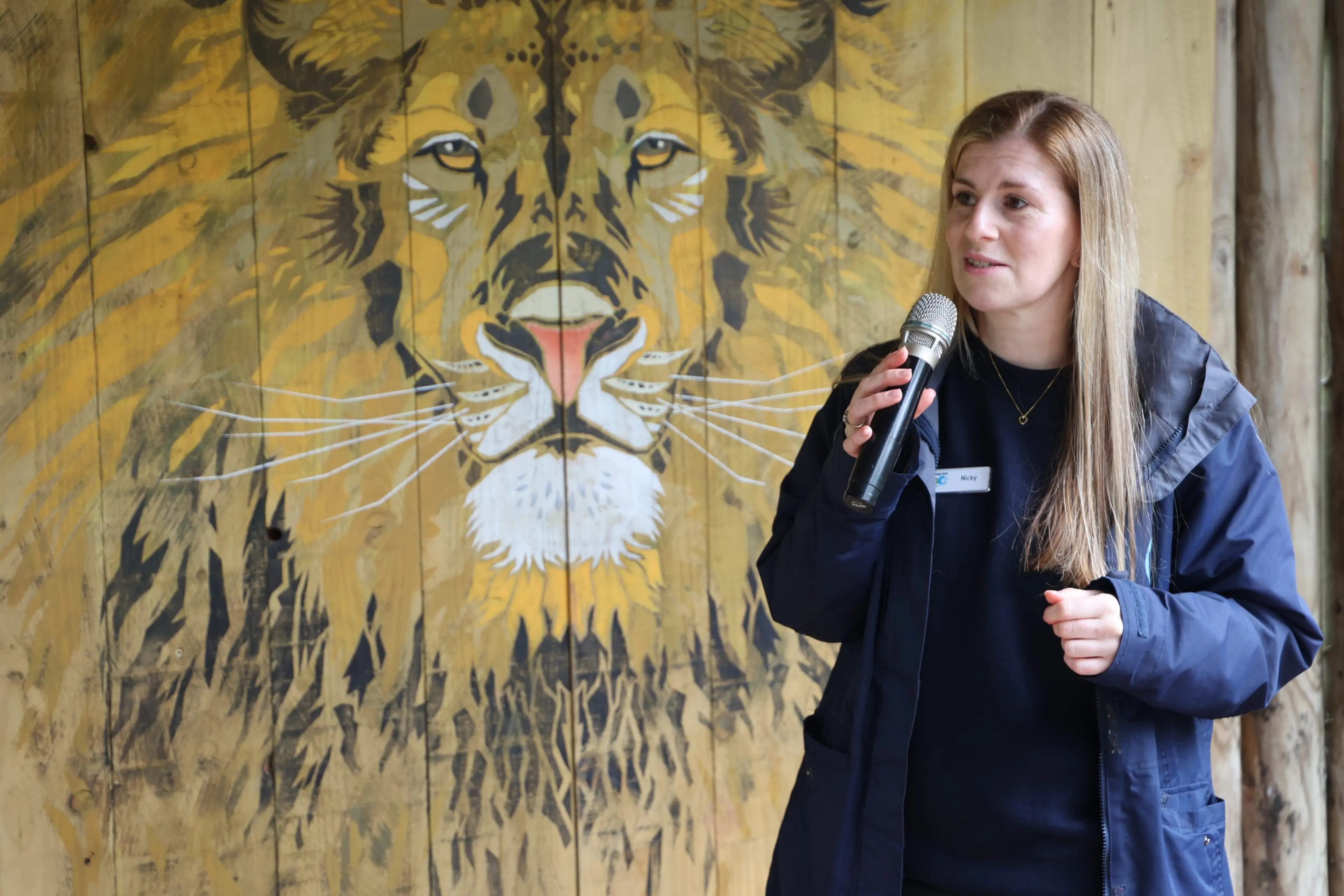 A woman speaks into a microphone in front of a large painted mural of a lion on a wooden wall.