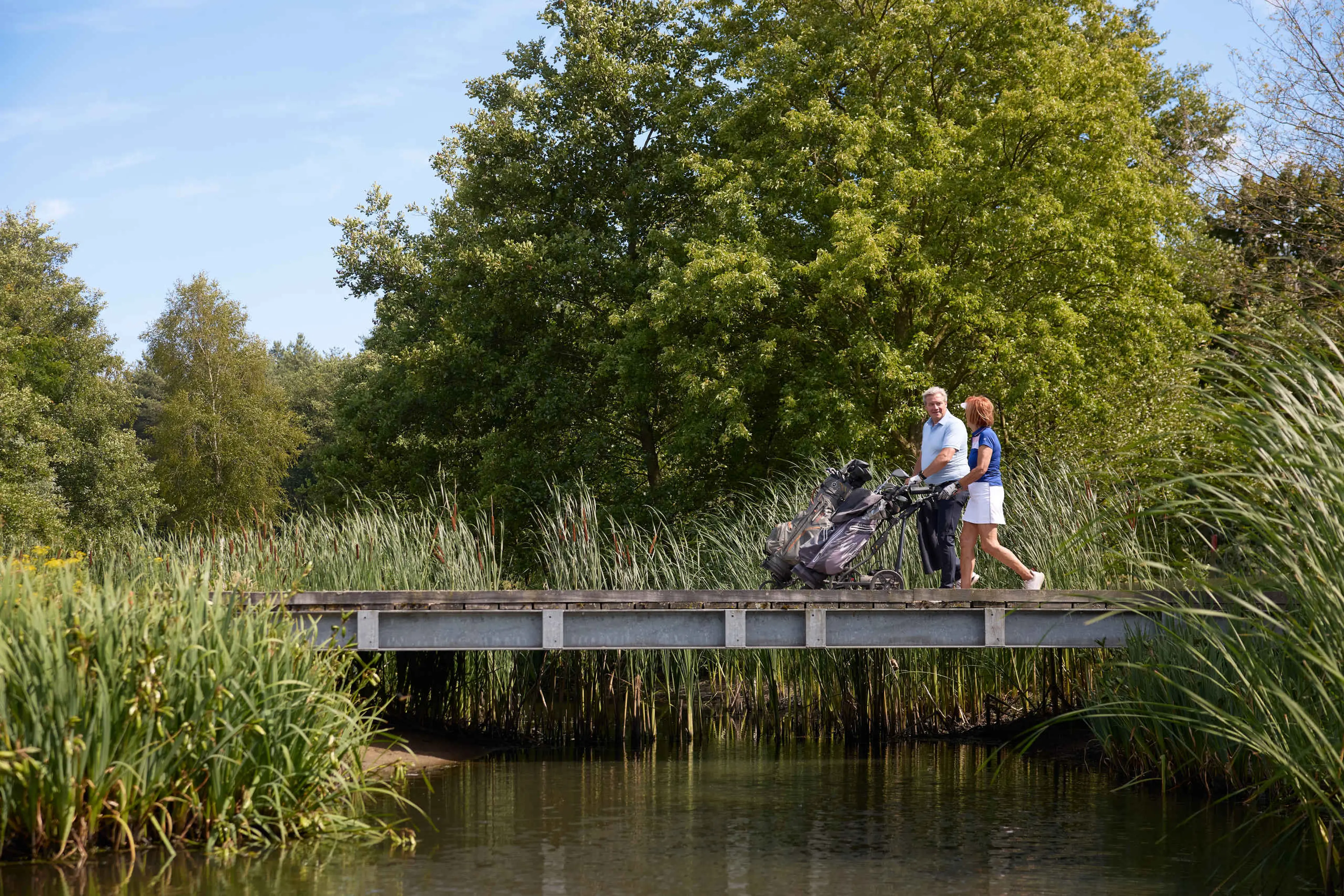 Een man en vrouw lopen met hun trolleys over de brug op de libellebaan bij Golf De Gulbergen