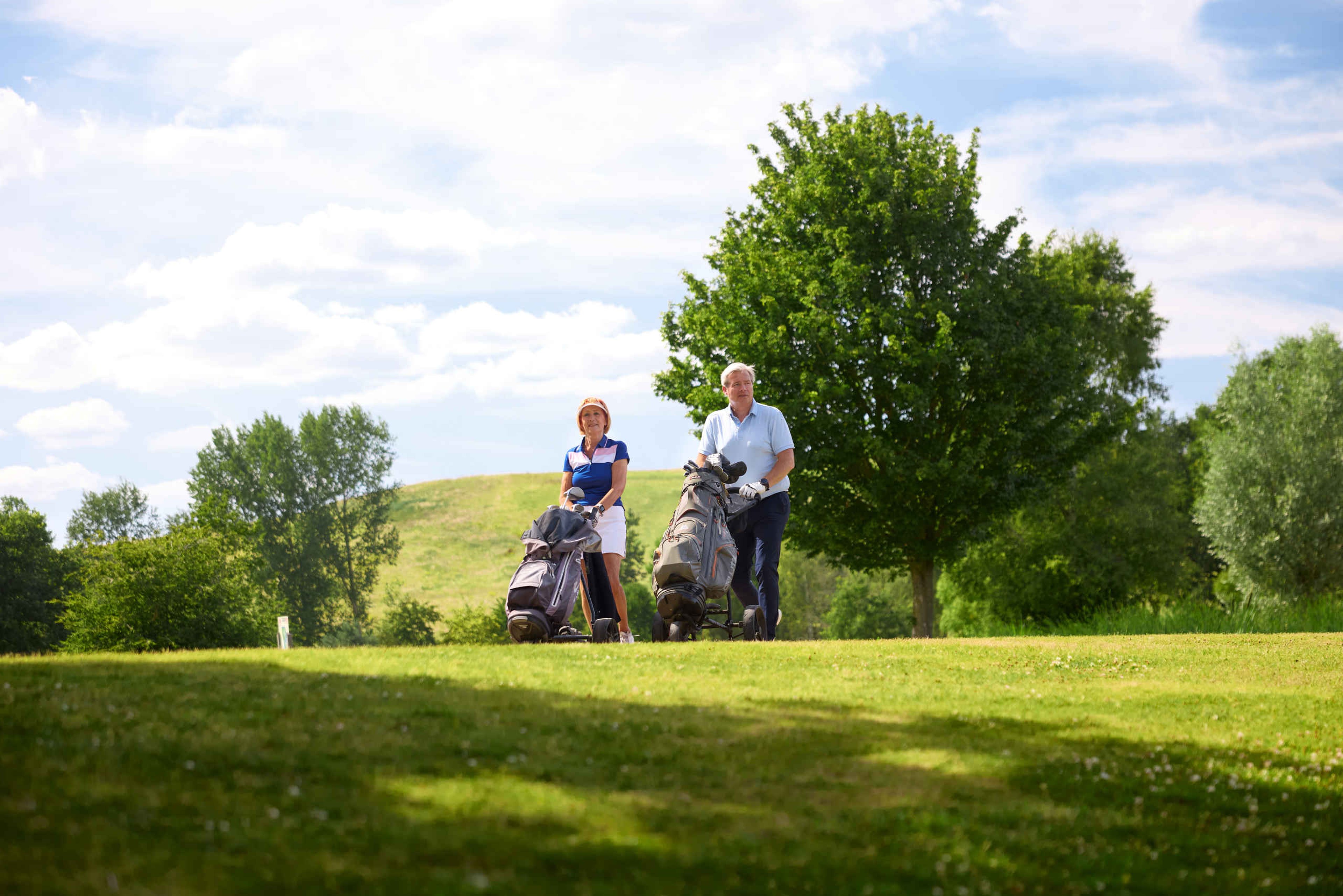 Een man en vrouw lopen met hun trolleys over de fairway van de libellebaan bij Golf De Gulbergen