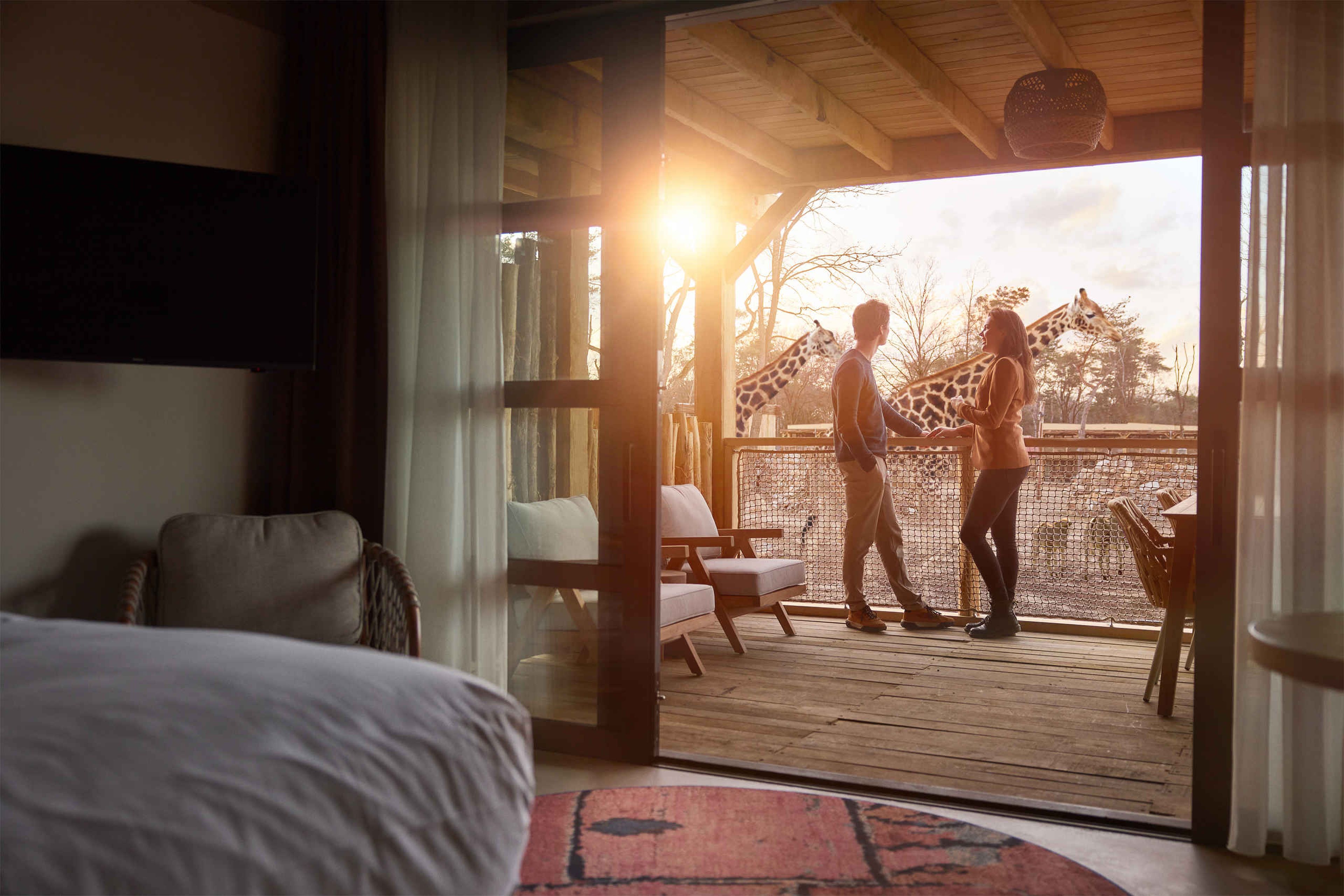 Een koppel staat op het balkon van een savanne room waar ze kijken naar giraffen op de savanne bij Safari Hotel Beekse Bergen, waar je kan overnachten na jouw bezoek aan Bar & Restaurant Nommos.