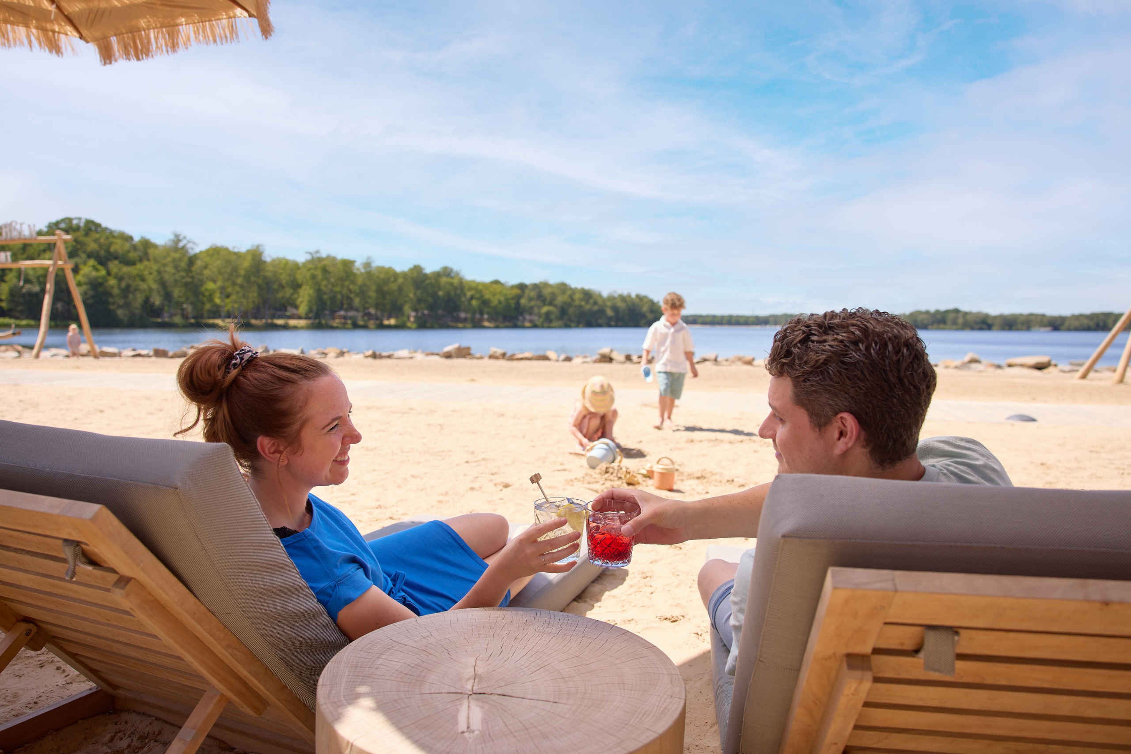 Een man en vrouw relaxen met een cocktail op een strandbedje terwijl hun kinderen spelen op het strand bij het Victoriameer bij Lake Resort Beekse Bergen.