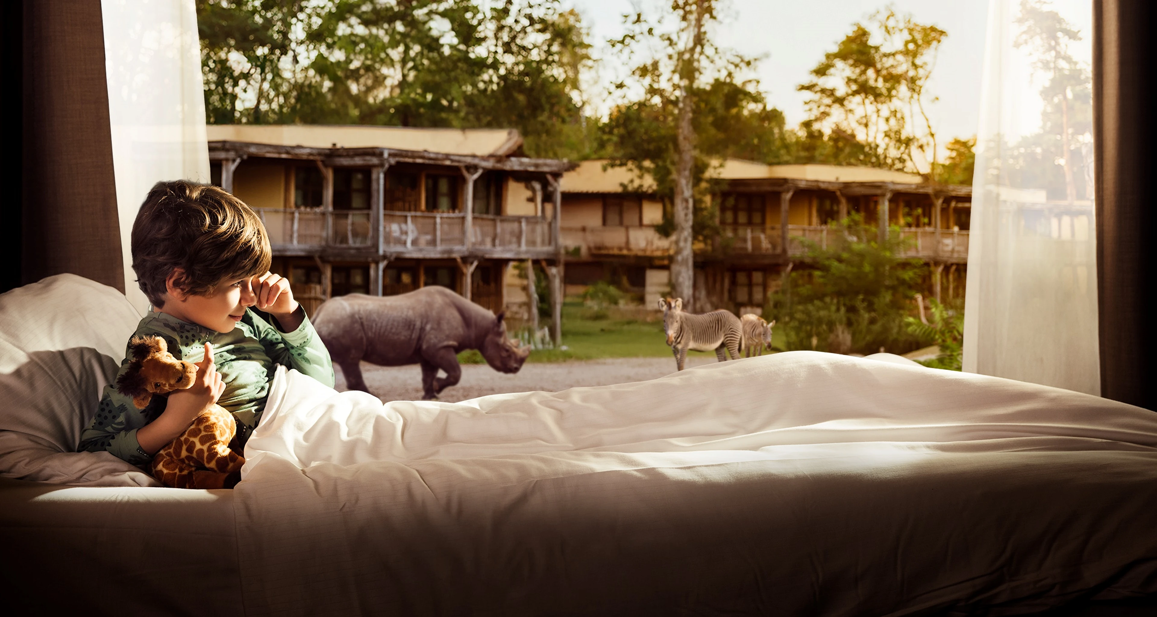 Een jongen ontwaakt in een Savanne Room met uitzicht op een savanne met giraffen, zebra's en neushoorns bij Safari Hotel Beekse Bergen.