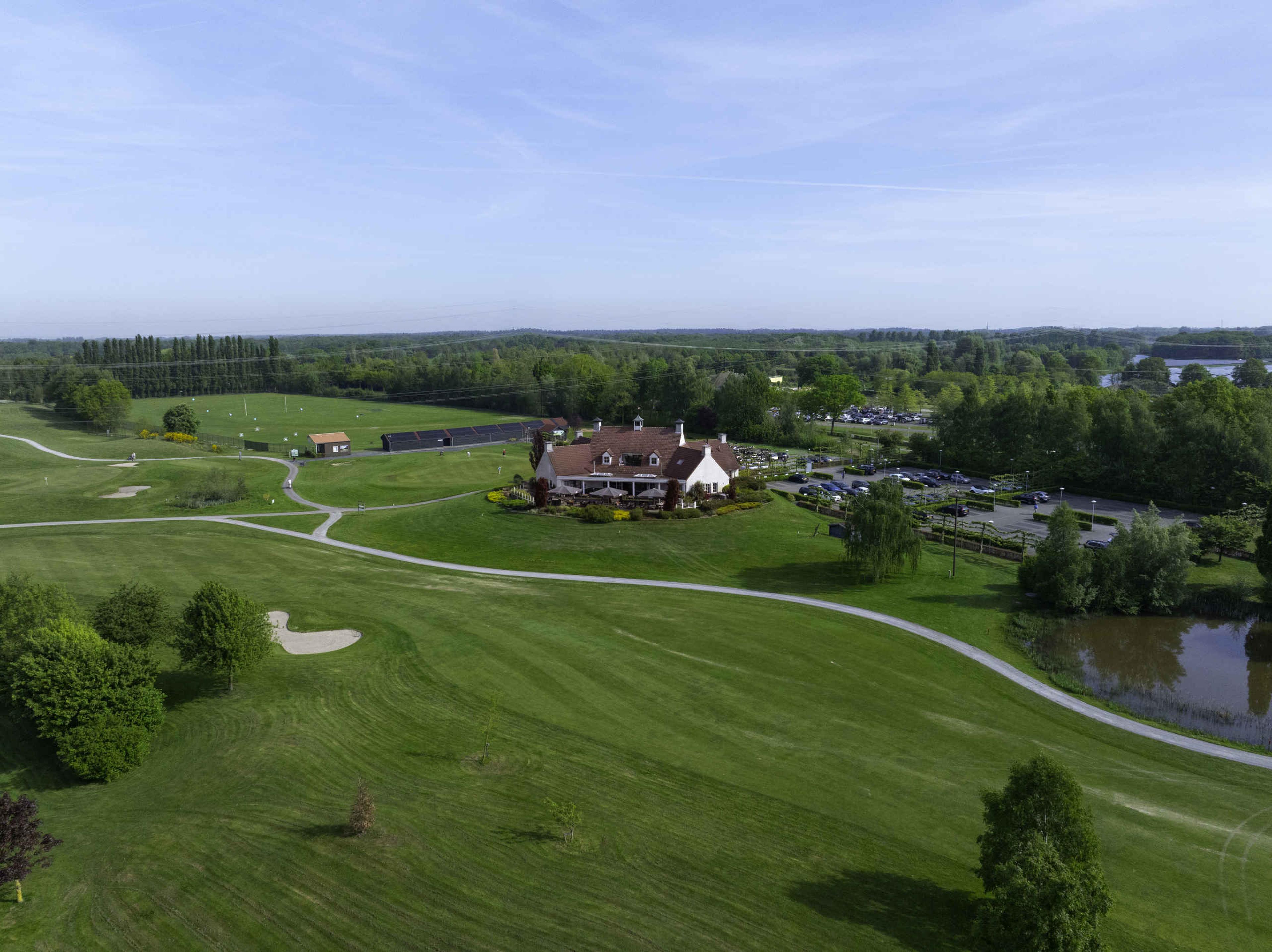 Een droneshot van de fairway van hole 9, het clubhuis, de putting green en de driving range van Golf De Gulbergen