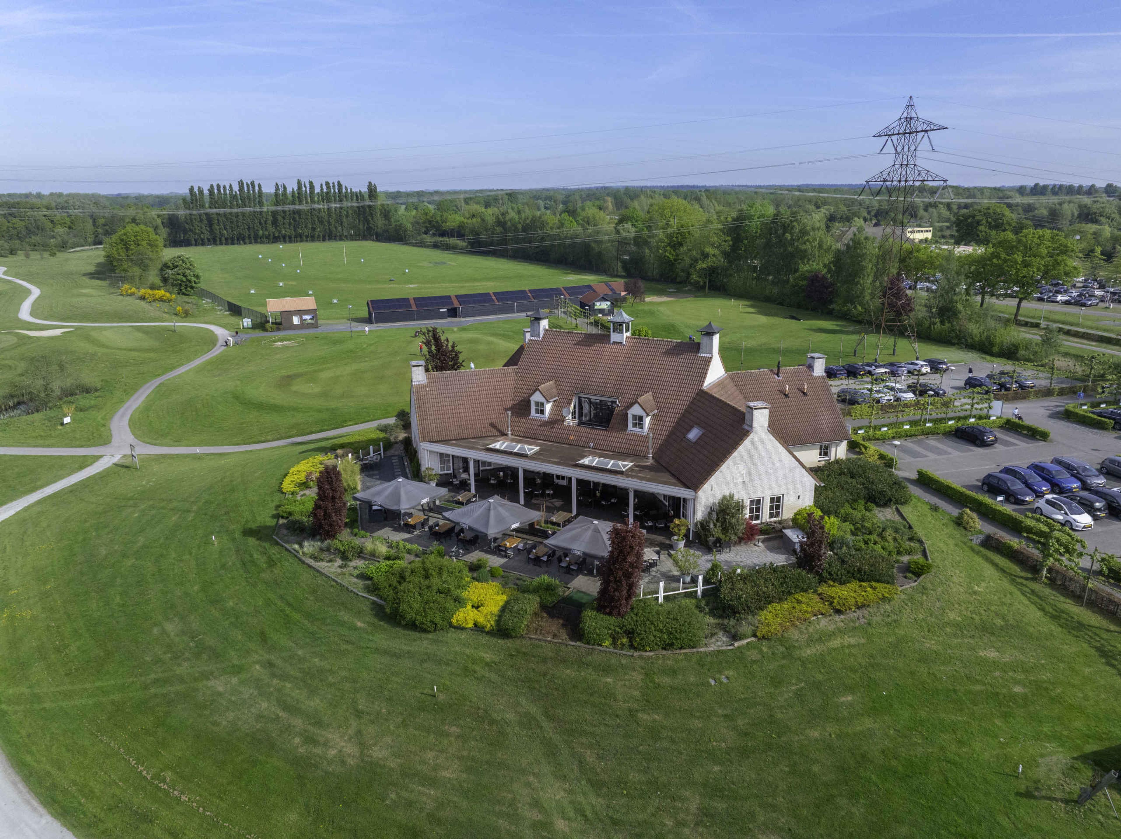 Een droneshot van het clubhuis met terras en restaurant en in de verte de driving range van Golf De Gulbergen