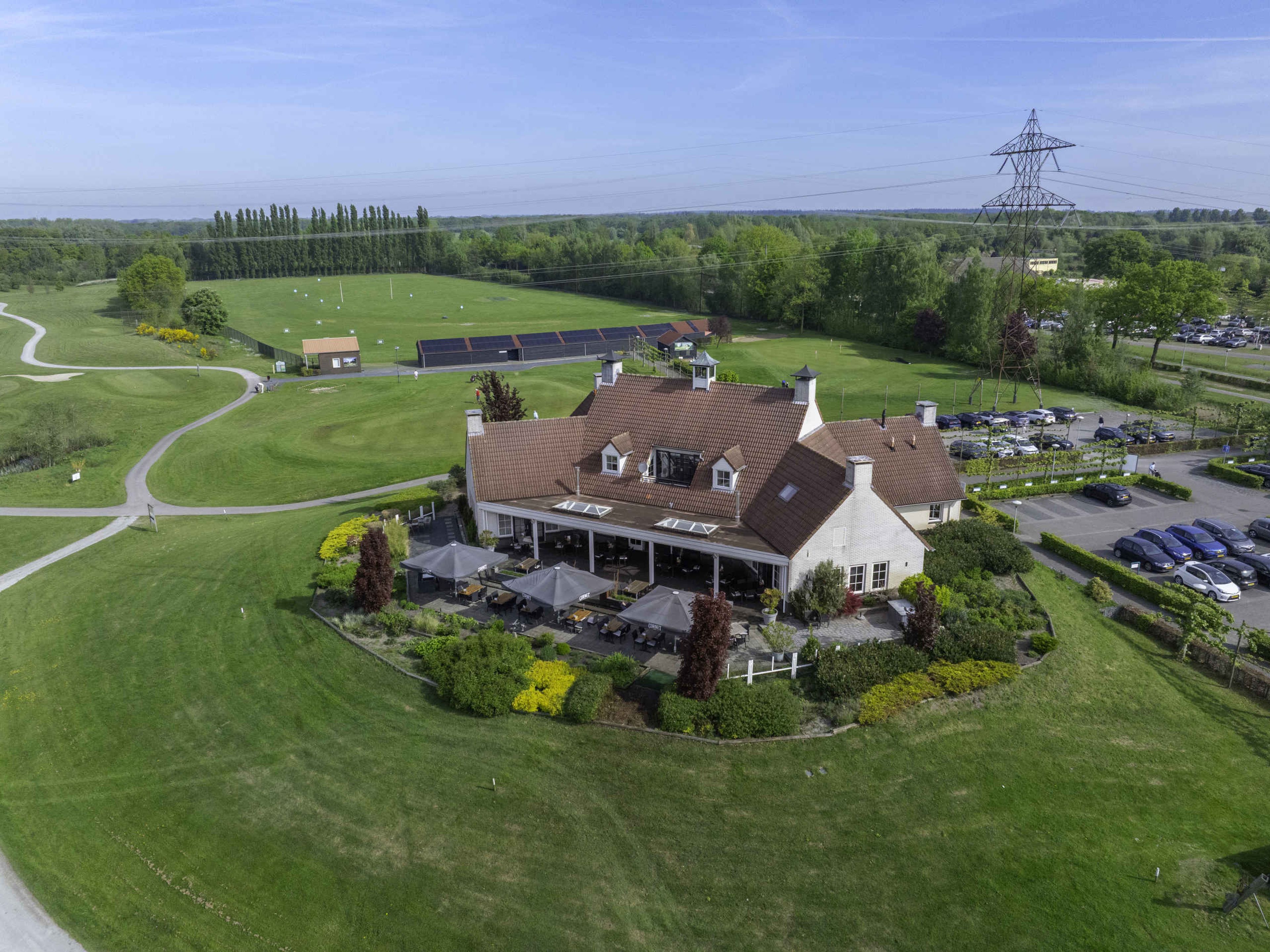 Een droneshot van het clubhuis met terras en restaurant en in de verte de driving range van Golf De Gulbergen