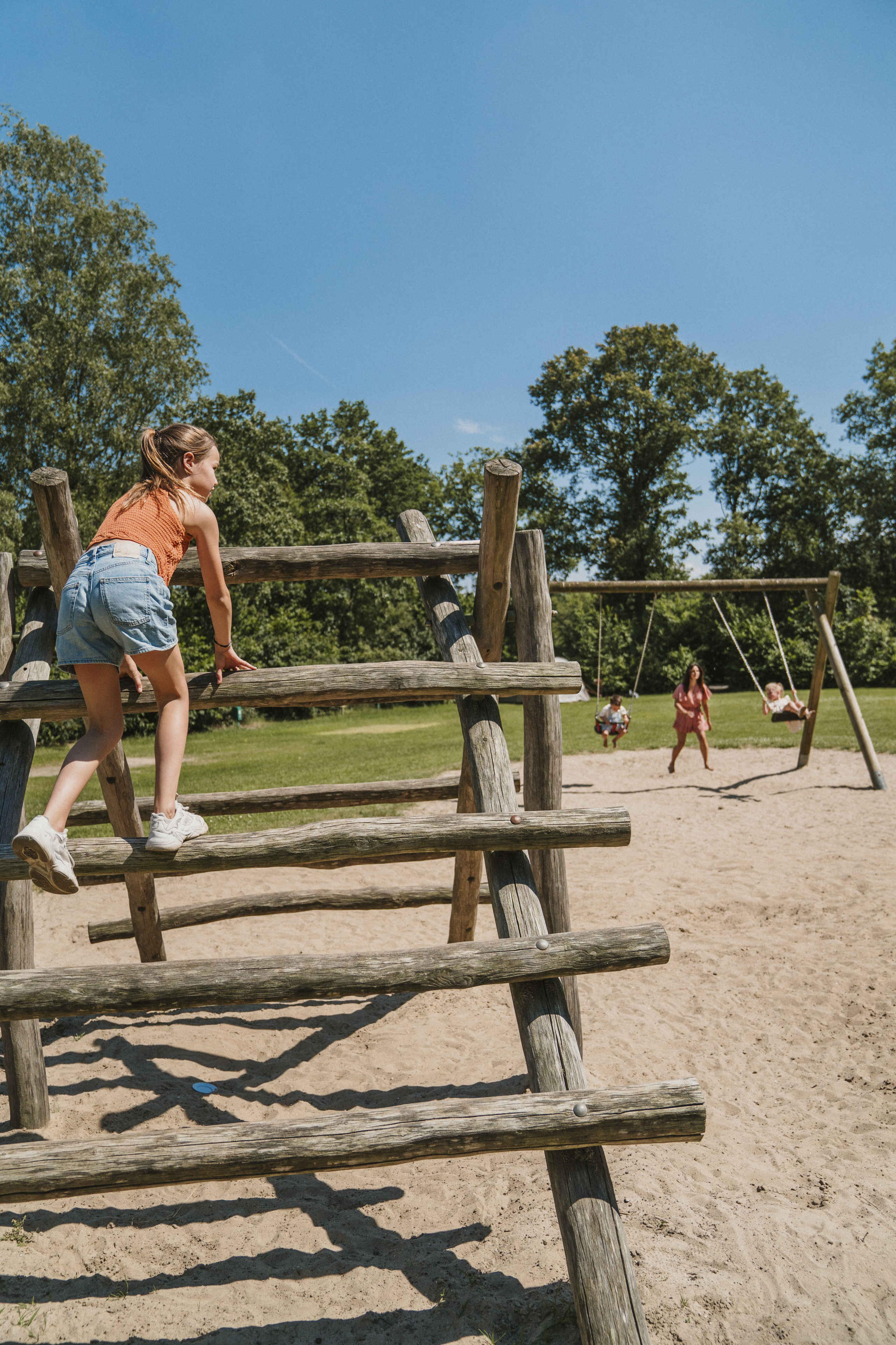 Een meisje klimt op een klimrek terwijl een gezin schommelt op de kampeerplaats bij Vakantiepark Dierenbos