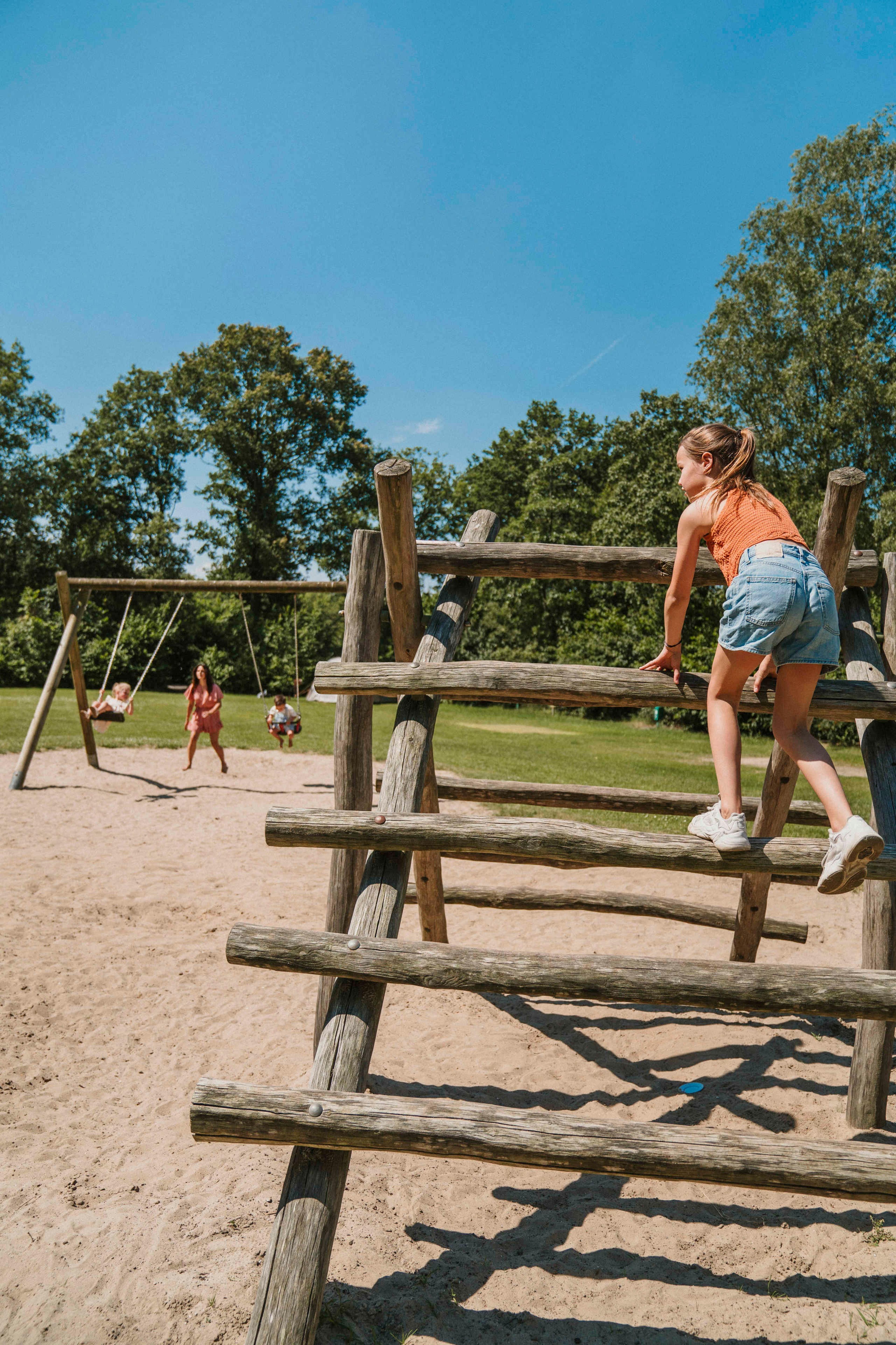 Een meisje speelt op een klimrek terwijl een moeder op de achtergrond met haar kinderen bij de schommels speelt bij Vakantiepark Dierenbos.