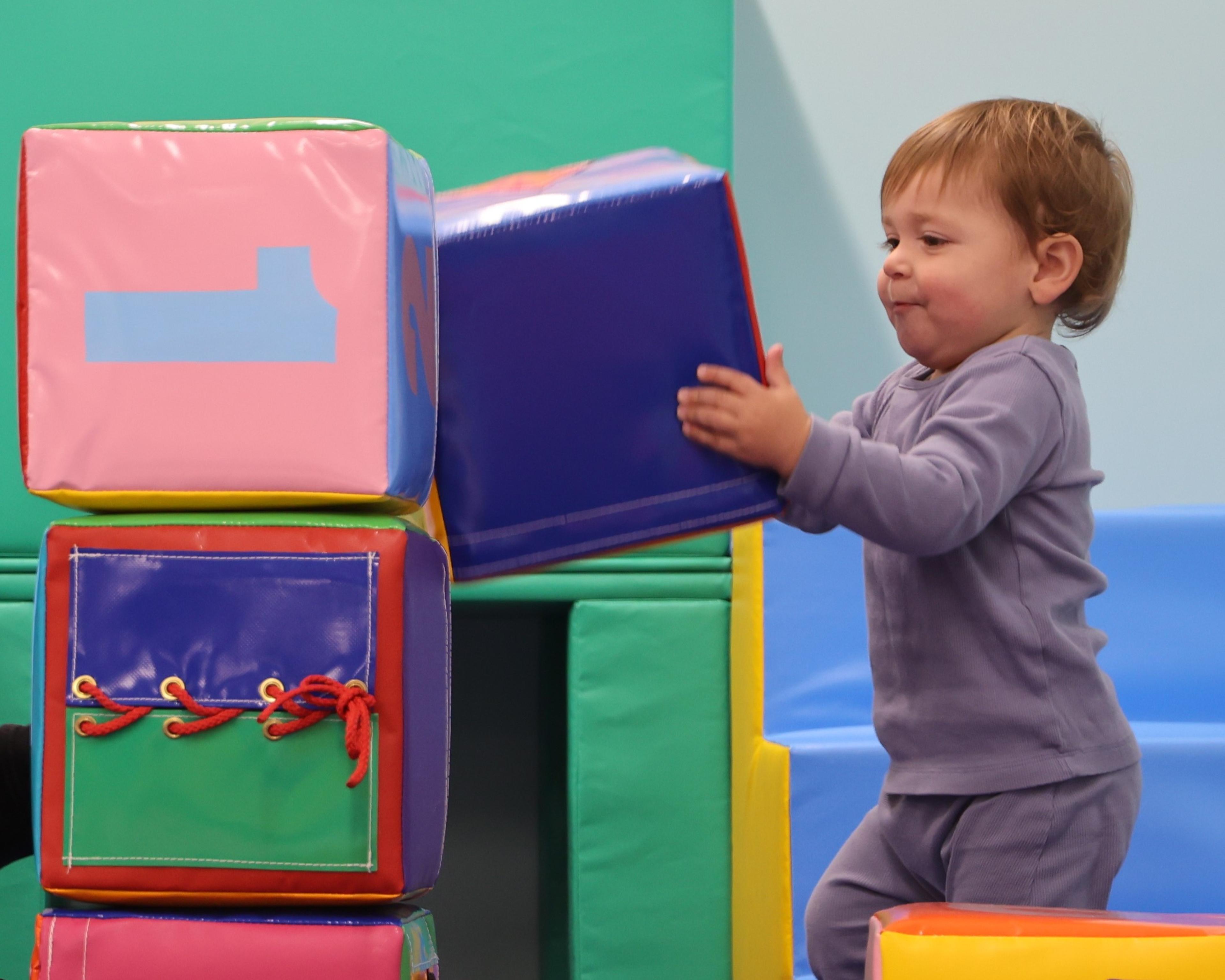 A toddler in a purple outfit is stacking colorful soft blocks indoors, concentrating on adding a large purple block to the stack.